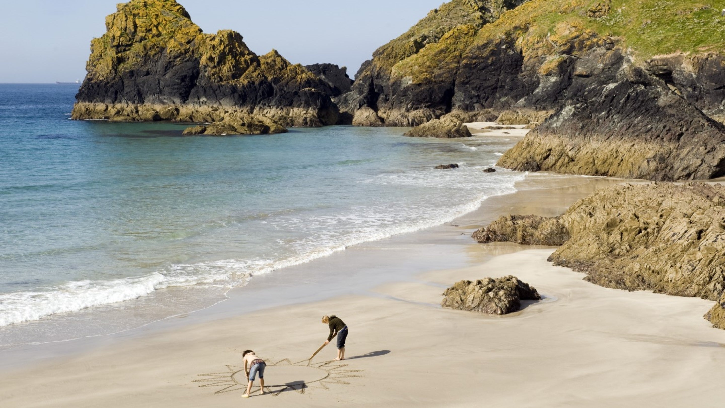 Two women drawing a huge sun on the sandy beach at Kynance Cove, Cornwall