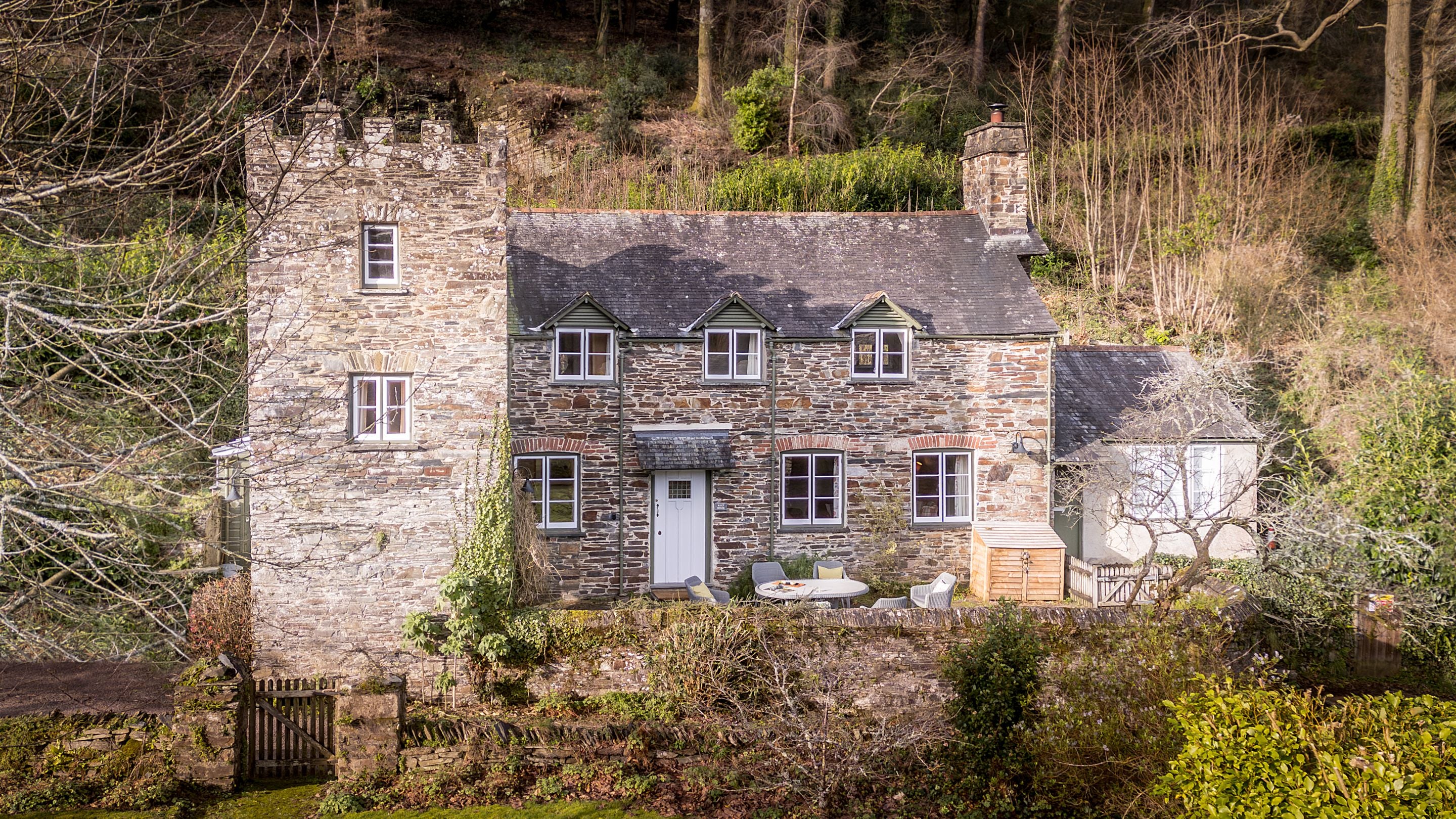 An aerial view of the exterior of Malt House, a stone cottage with a small tower, patio for outdoor dining and private garden, Cornwall