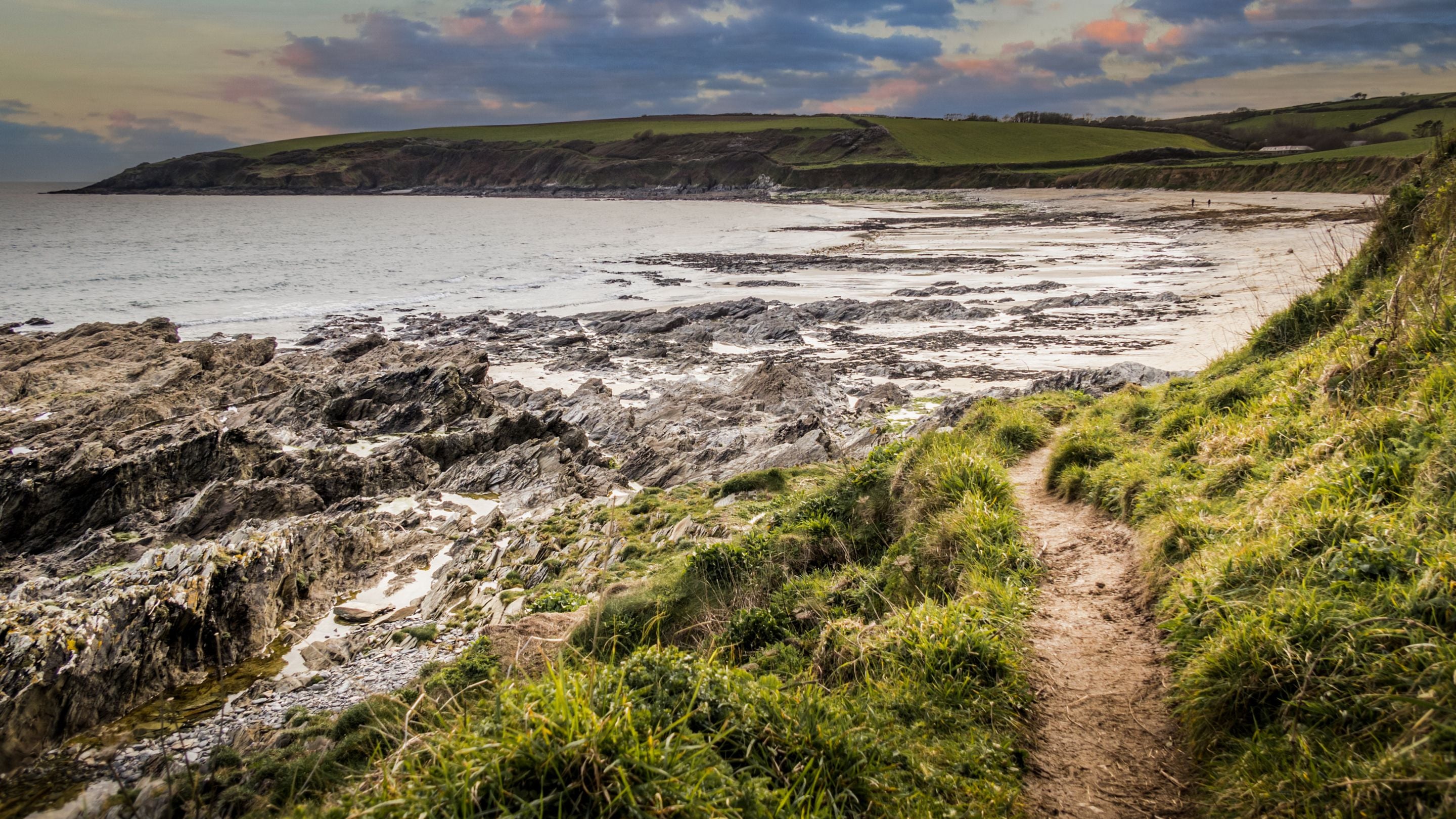The path leading to sandy Towan Beach, with rockpools in the foreground and green fields above the beach in the background, Cornwall