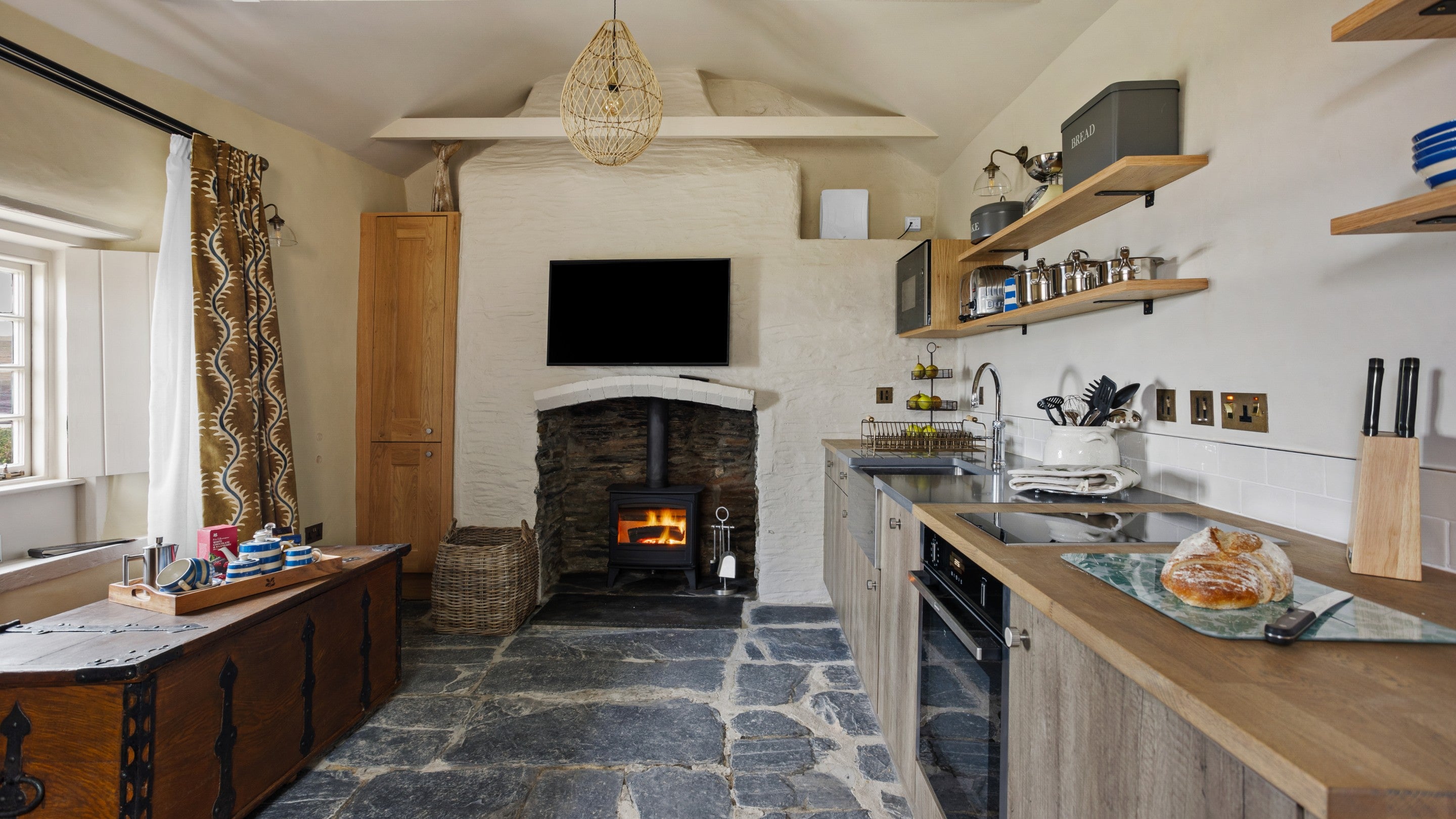 A view of the living space at Tom Parsons' Hut, showing the woodburner with television above the fireplace, kitchen and dining table, Cornwall