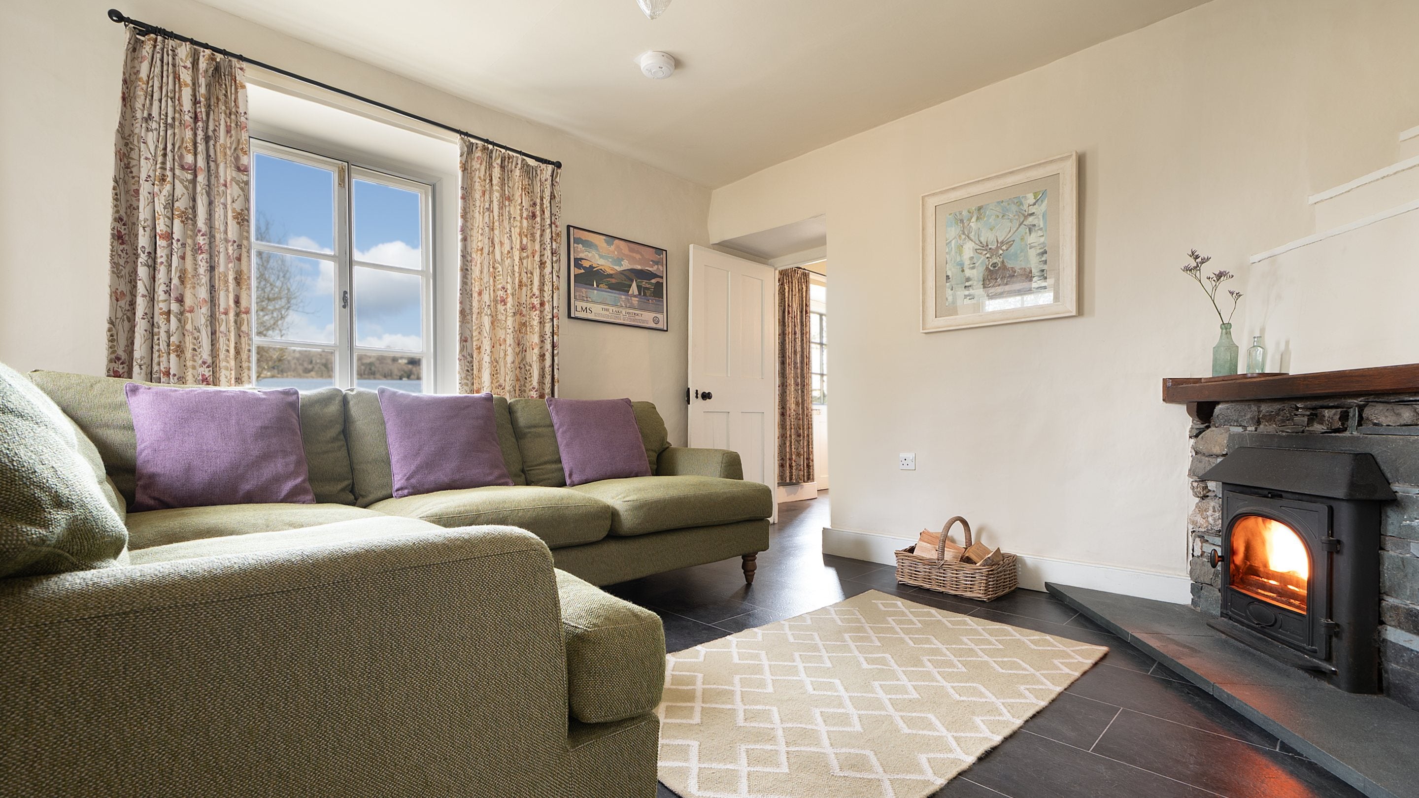 The sitting room at High Strawberry Gardens with corner sofa, woodburner and view of Lake Windermere, Cumbria