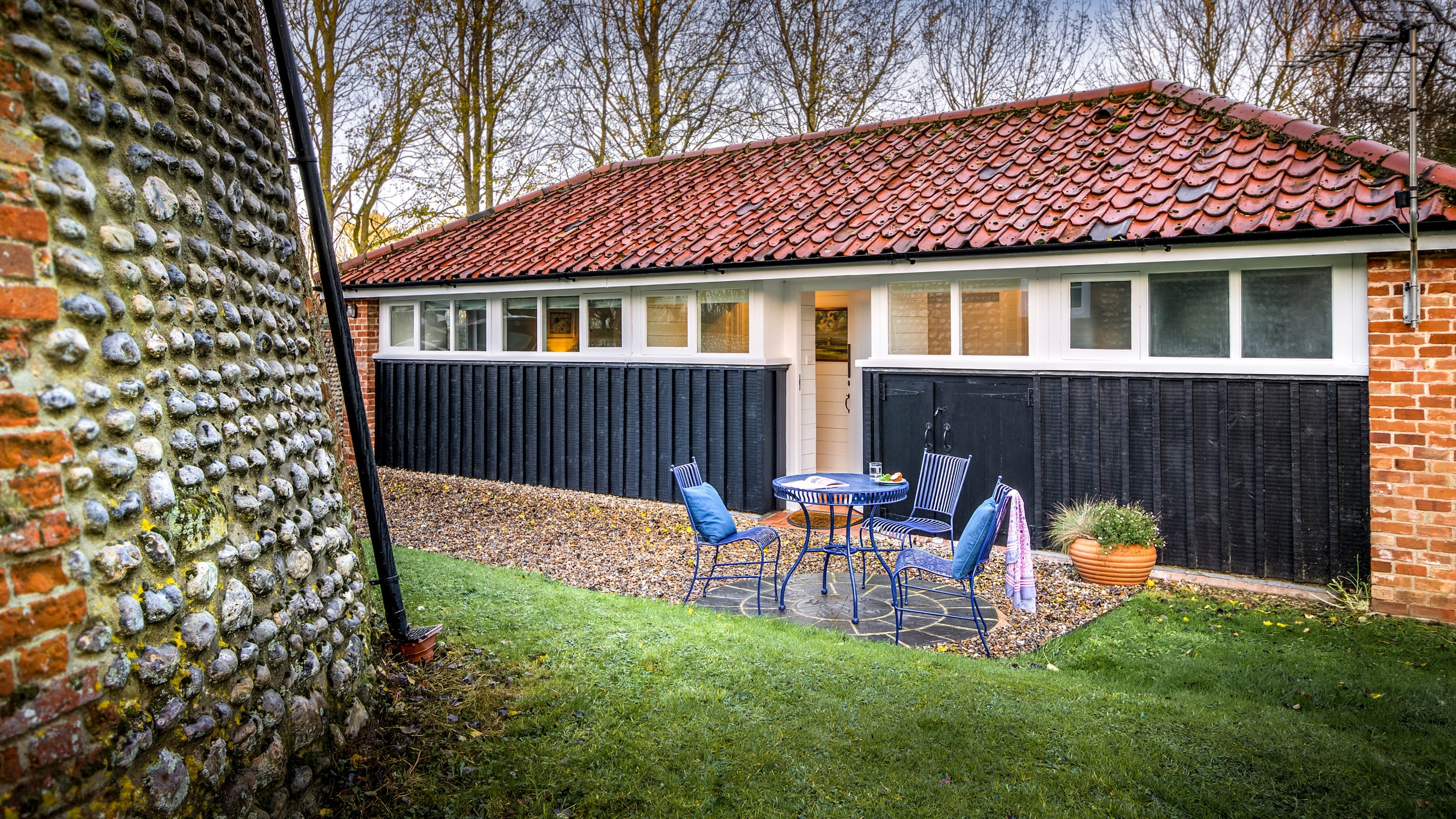 The exterior of Blakeney Lodge and its walled garden, with a patio with outdoor dining furniture, a lawn and the base of the windmill, Norfolk