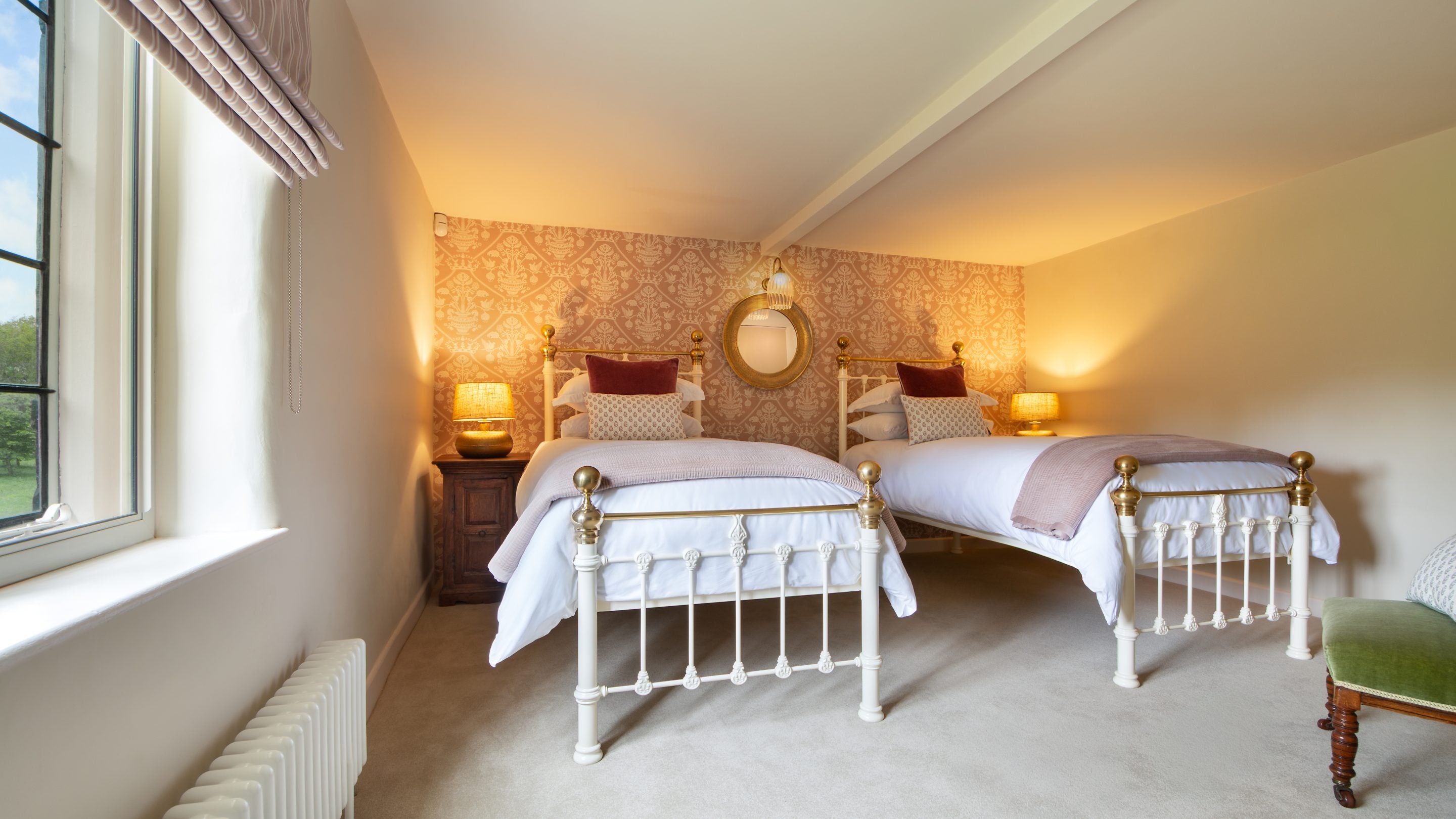 A first-floor bedroom with twin beds with white and gold metal bed frames, each with a bedside table and lamp, a wallpapered wall in a pink and white pattern with a round mirror, one green velvet chair and a window looking over the parkland, at Blickling Tower, Norfolk