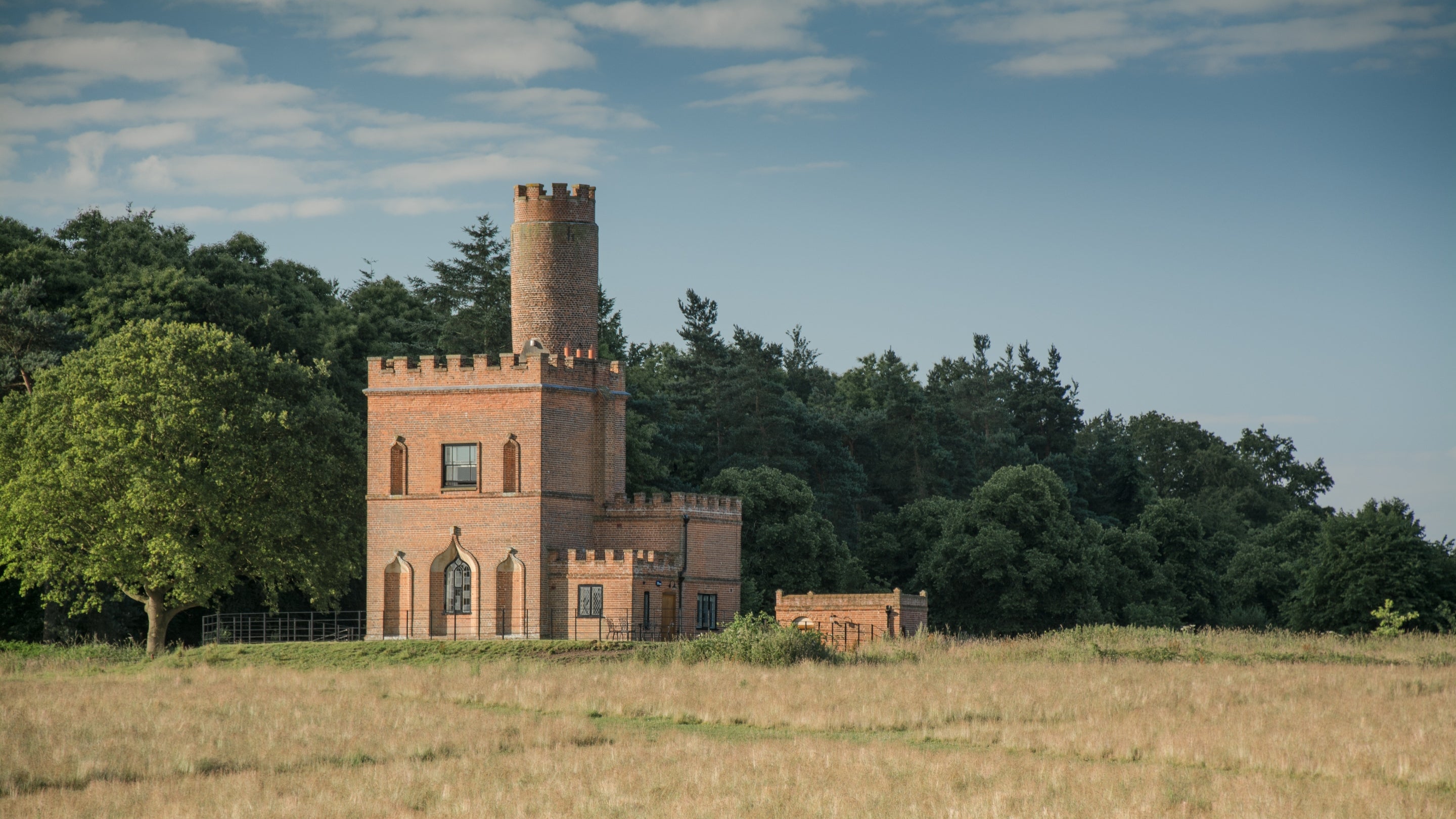 The exterior of the red-bricked Blickling Tower with it's square base and cylindrical tower rising out of the top. The holiday cottage is viewed on a sunny summers day with a wooded landscape right behind it.