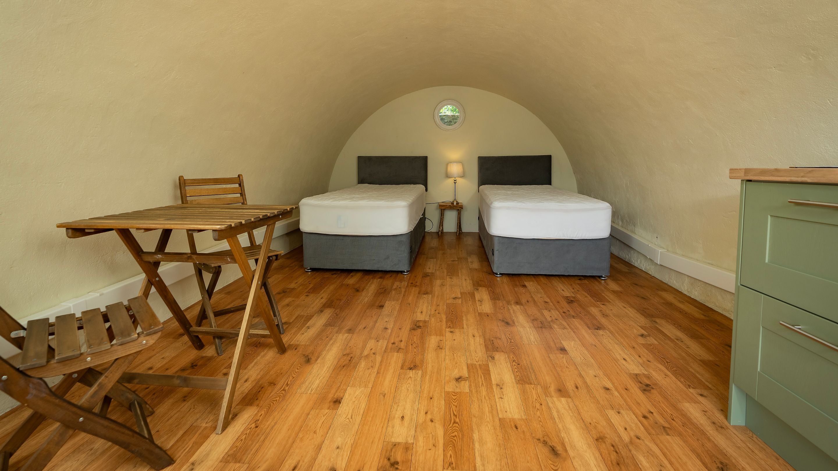 Inside one of the pods at Crom, showing the two single beds with mattresses but no bedding, a table and two chairs and kitchen drawers