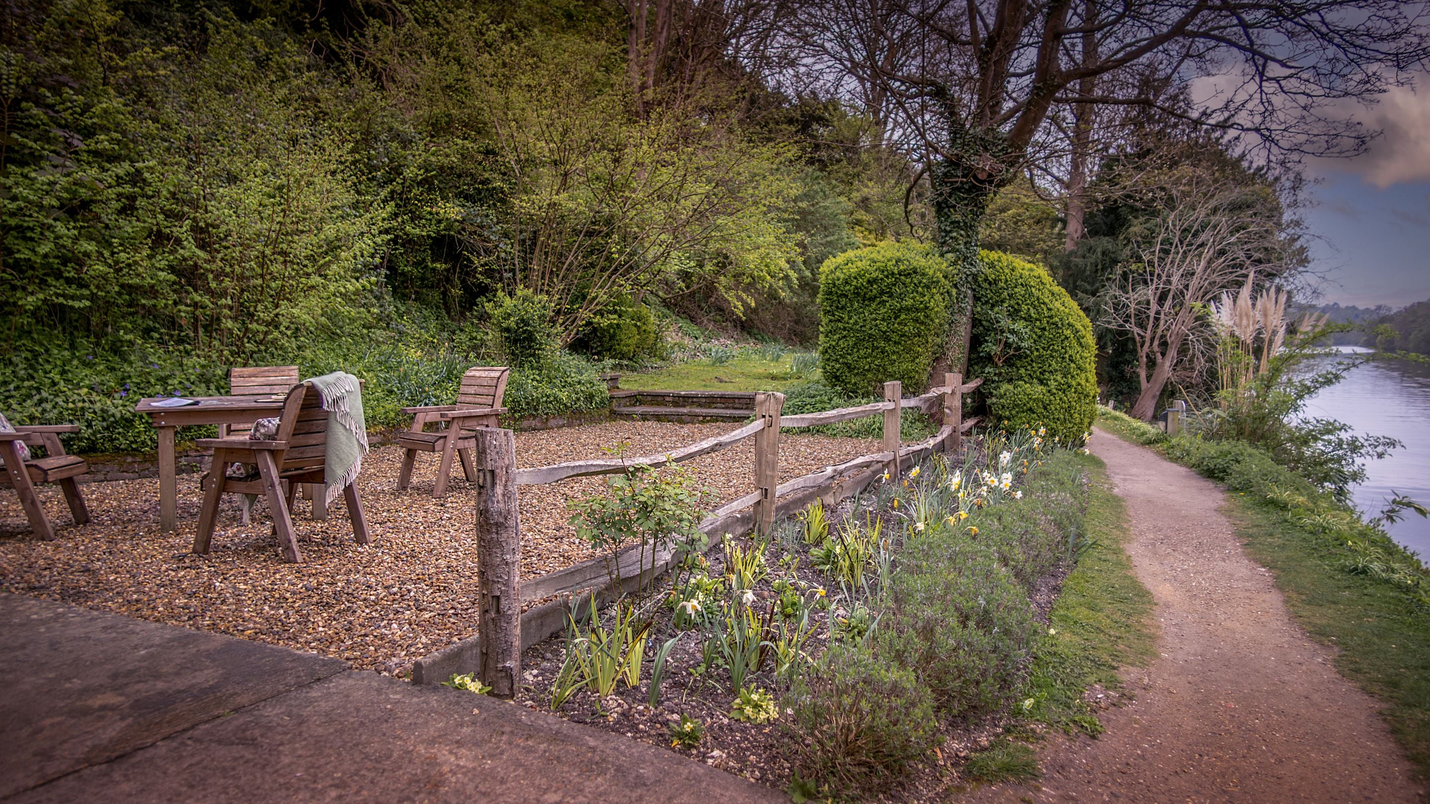 The garden at Cliveden Ferry Cottage, with gravelled area with dining furniture and small grassed area, next to the footpath on the bank of the River Thames, Berkshire