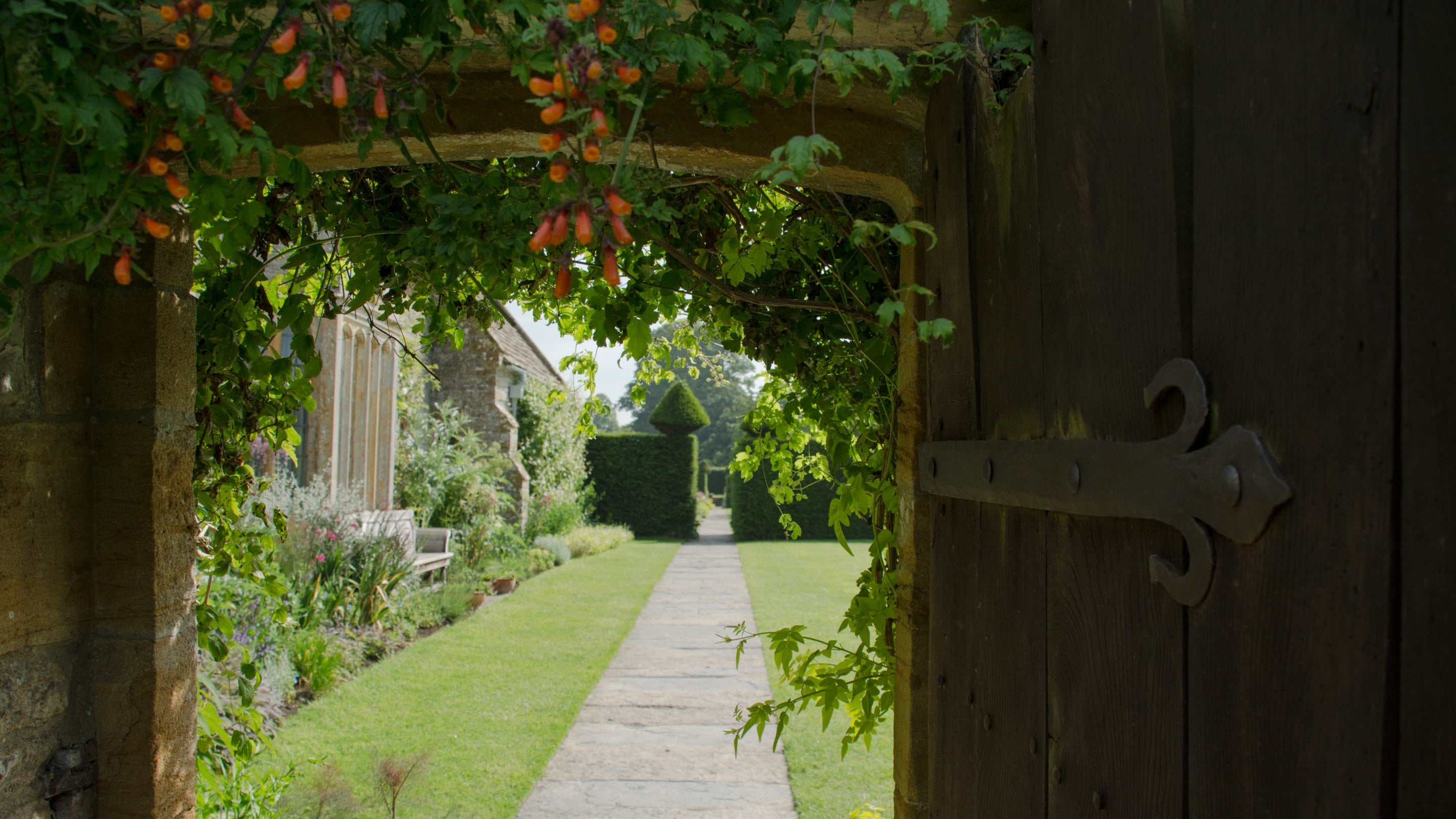 Part of the garden at Lytes Carey Manor, with paved stone path passing the manor, flower beds and a sculptured hedge, viewed from an old wooden door, Somerset