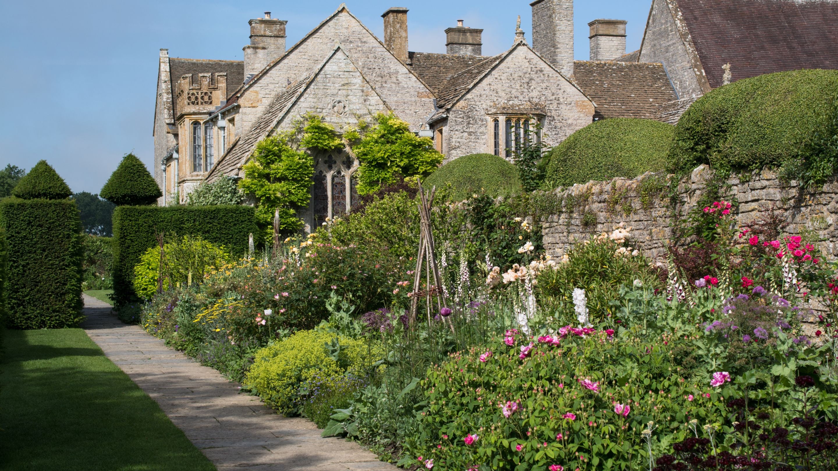 Part of the garden at Lytes Cary Manor, with a path past an old stone wall, flowerbeds, decorative hedges and the manor in the background, Somerset