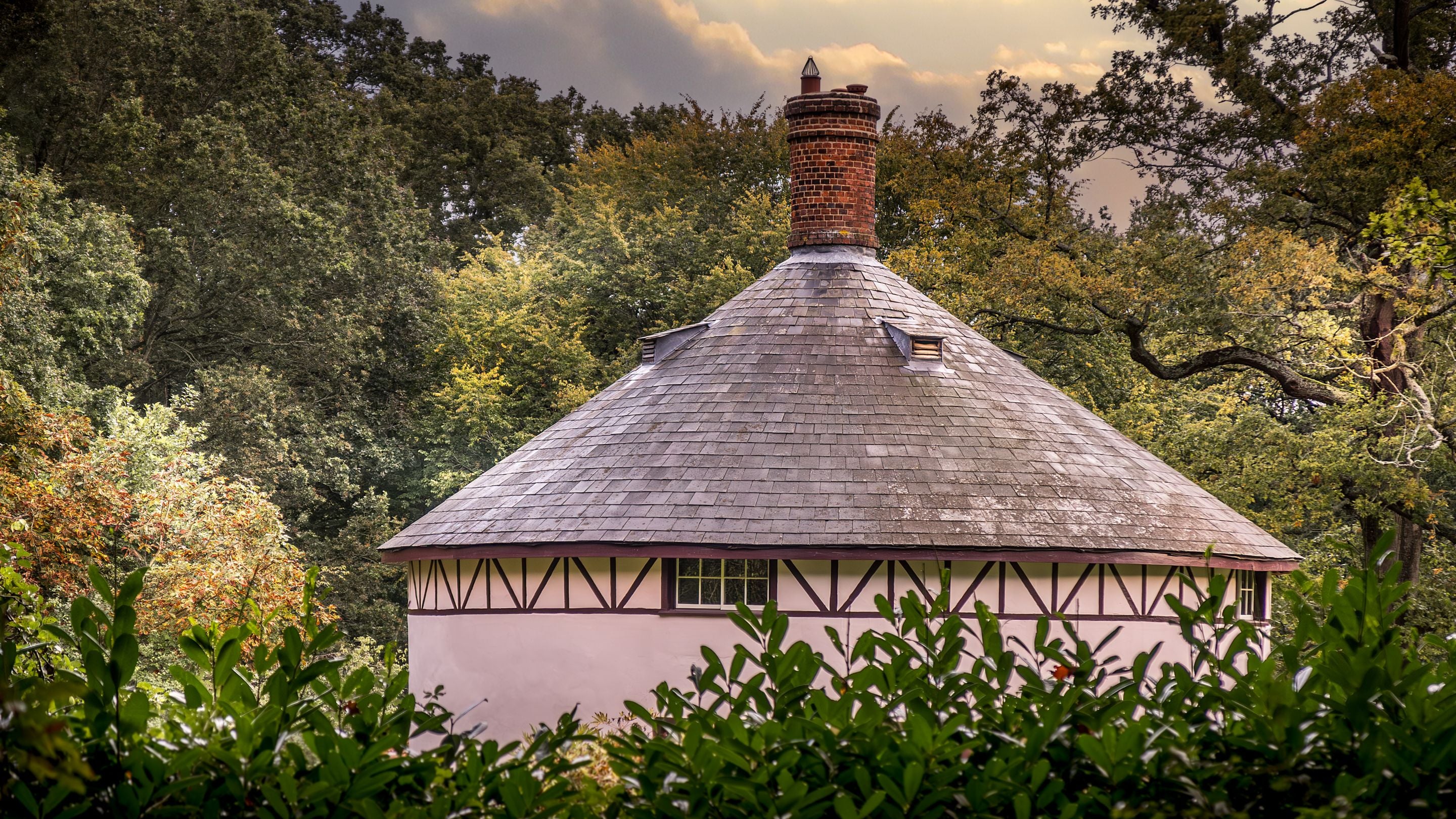 Looking through the woodland at the conical roof of The Round House, with cylindrical, red-bricked chimney in the centre, Suffolk
