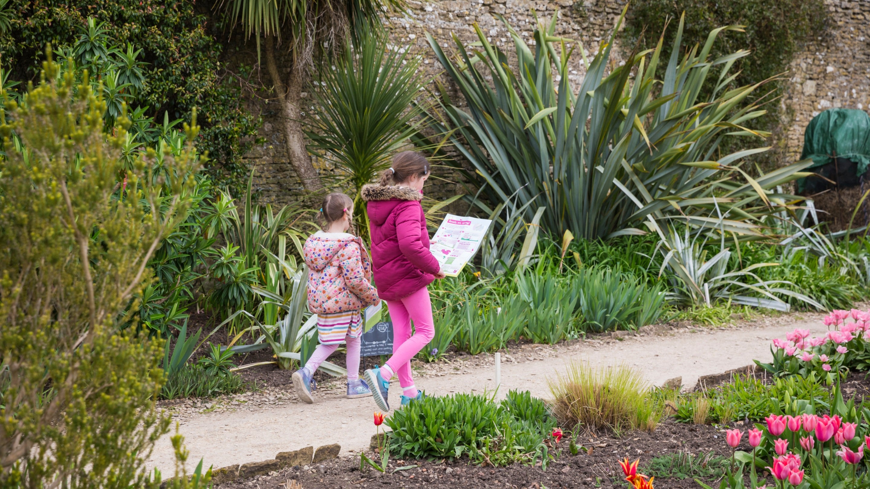 Two young girls walking along path in garden looking at Easter trail packs