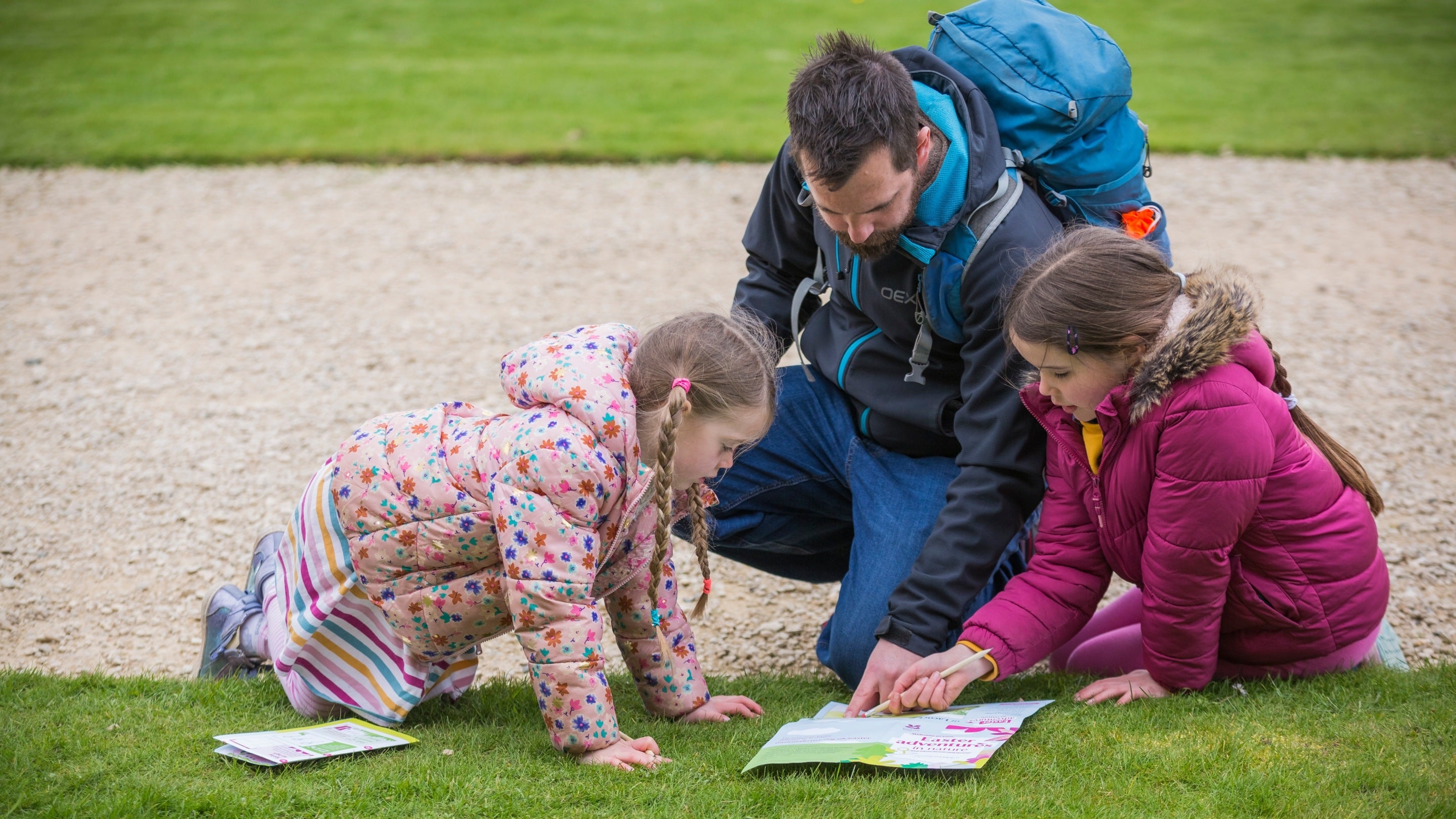 An adult and two children sit on the grass by a path doing an Easter trail at Lacock Abbey, Wiltshire