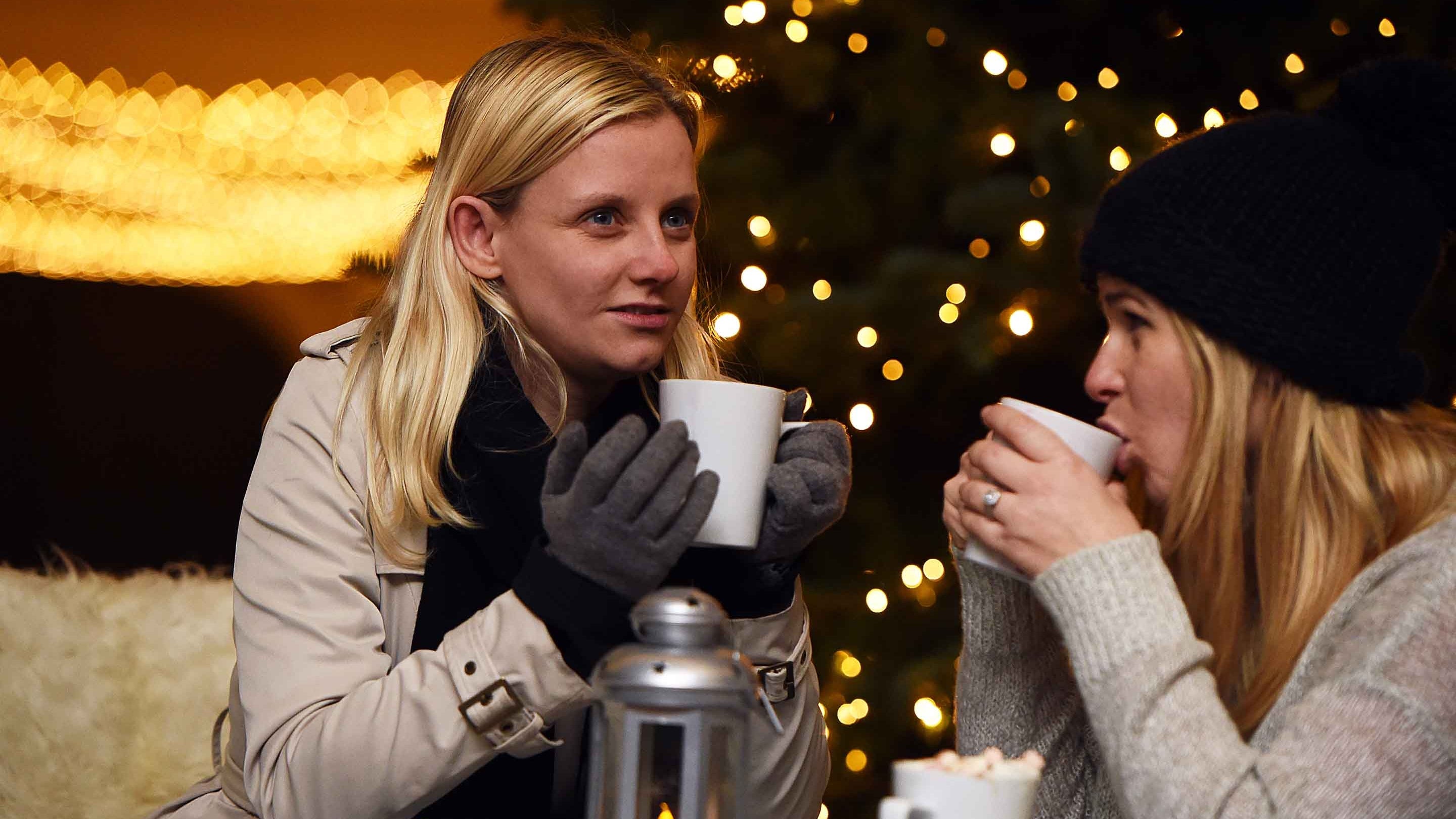 Two visitors drink hot drinks while sat outside a cafe with Christmas lights in the background
