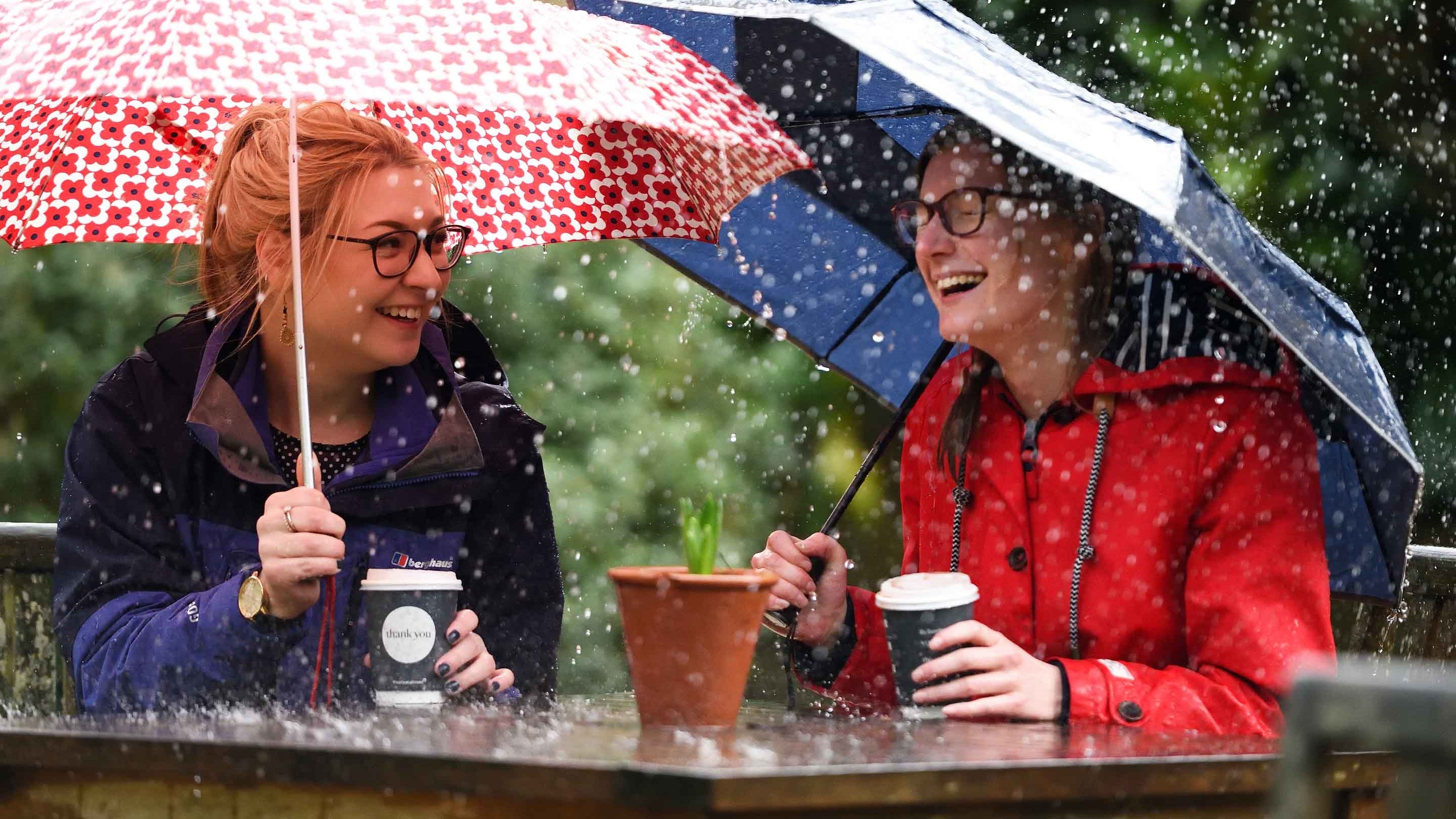 Two women sit laughing with takeaway cups and holding umbrellas at a table outside at Dyrham Park, Gloucestershire, on a rainy day.