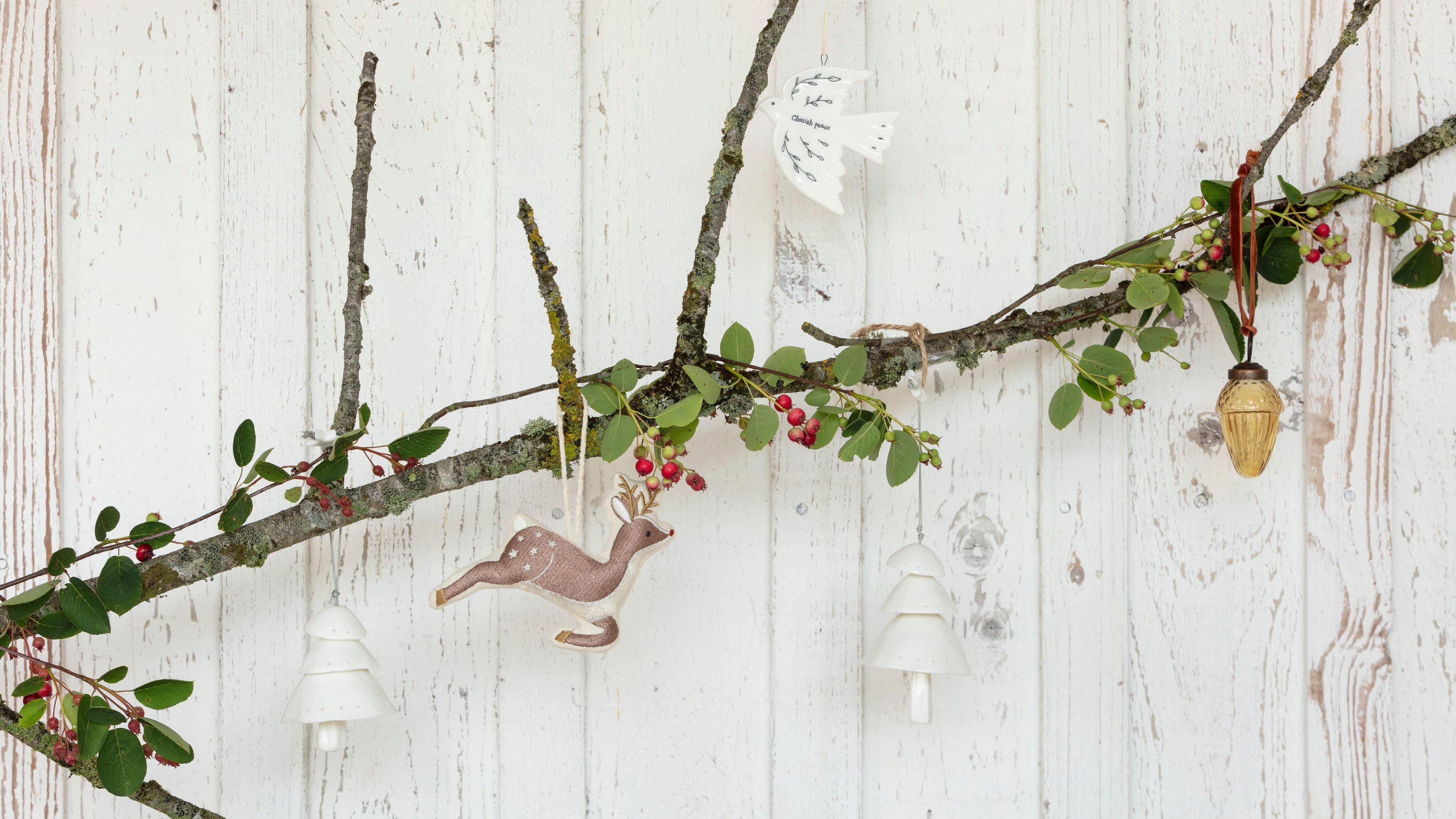 A selection of Christmas decorations including two white ceramic trees, a reindeer, a white ceramic dove and a gold acorn hung from a twig with winter foliage.