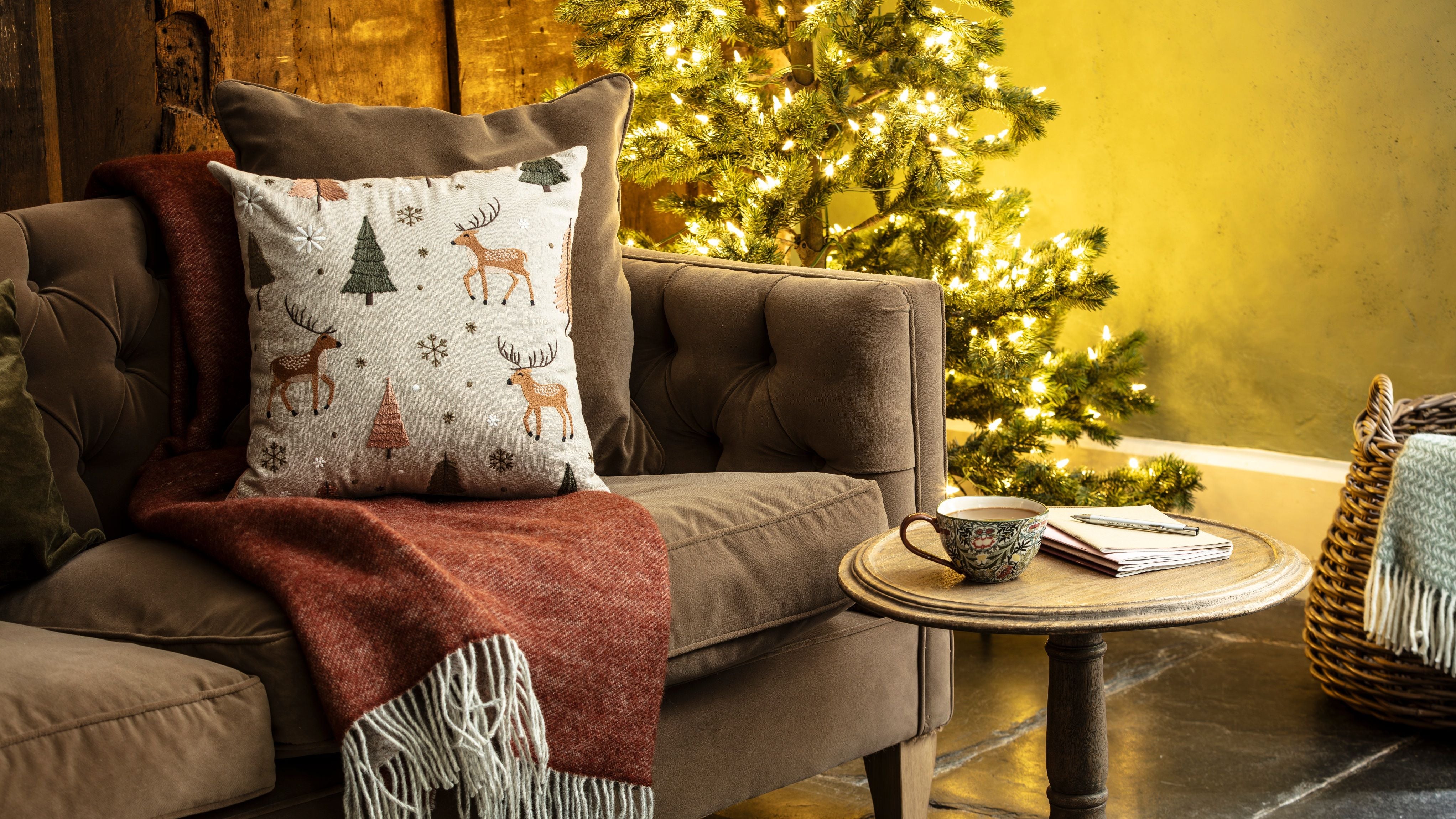 A living room with a lit Christmas tree. On the sofa is a terracotta-coloured throw with a cushion decorated in a pattern of reindeer and Christmas trees.