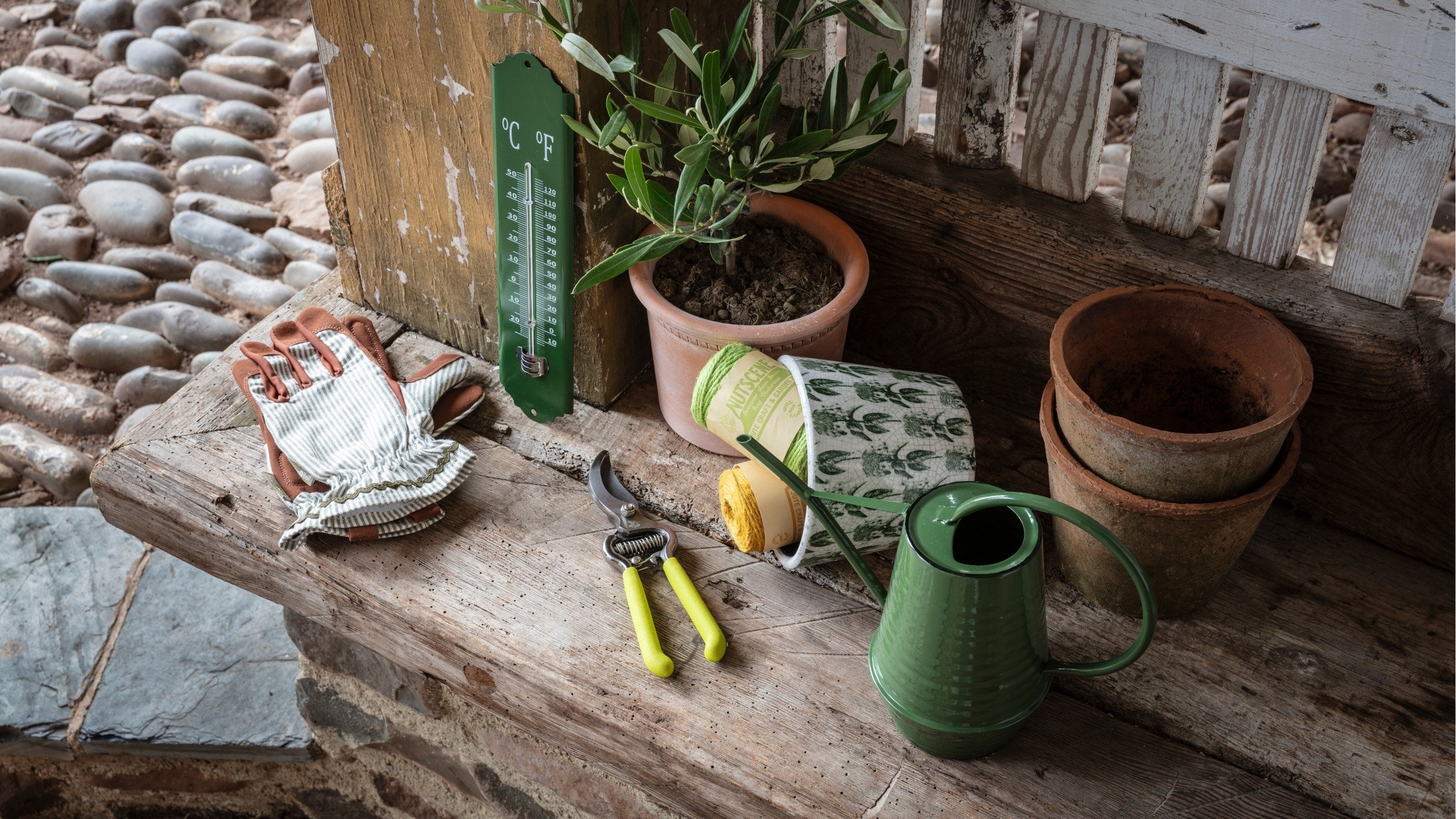 A selection of gardening accessories including gloves, secateurs, a thermometer, a watering can and a ceramic pot with balls of twin inside.