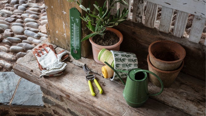 A selection of gardening accessories including gloves, secateurs, a thermometer, a watering can and a ceramic pot with balls of twin inside