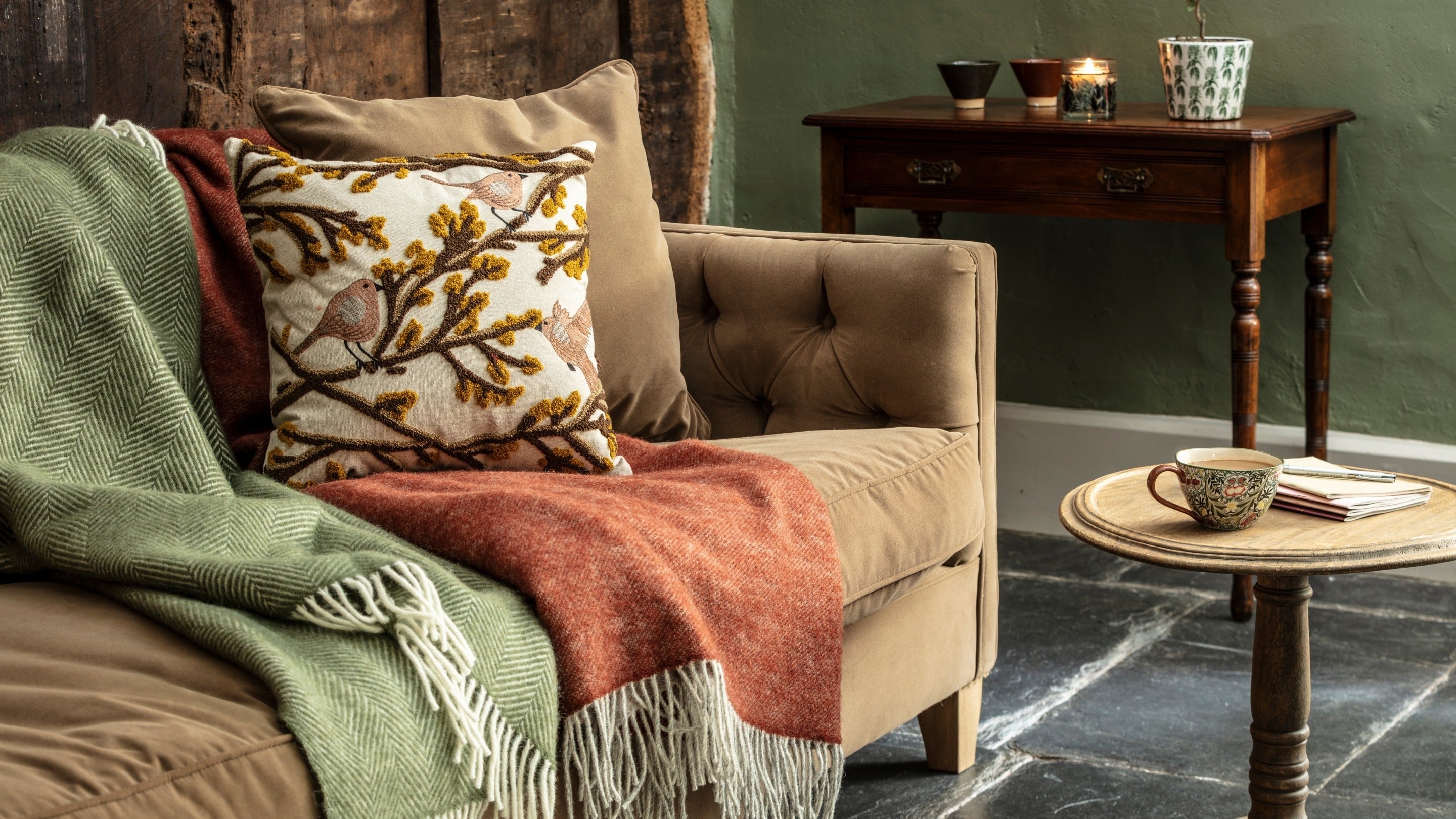 Throws and a cushion in autumnal colours on a sofa, with homeware items including a mug, plant pot and small decorative bowls in the background.