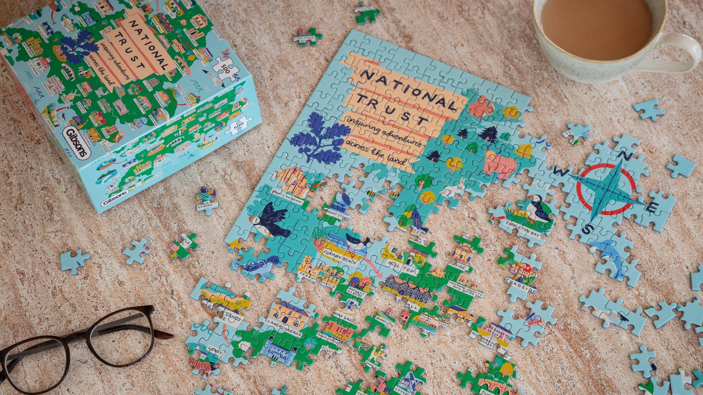 A half-finished puzzle of National Trust places on a table, with a cup of tea and a pair of glasses