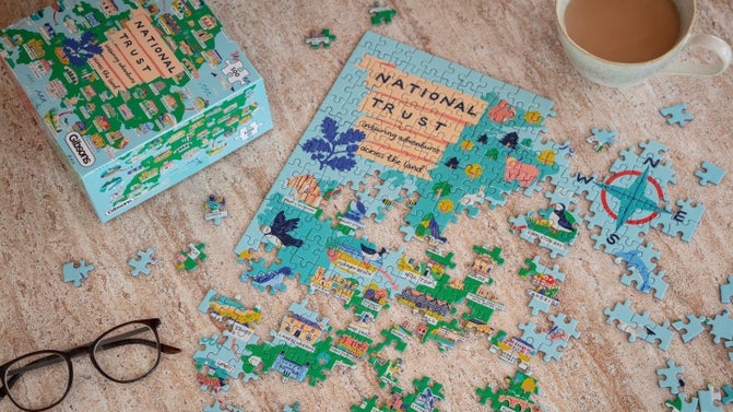A half-finished puzzle of National Trust places on a table, with a cup of tea and a pair of glasses.