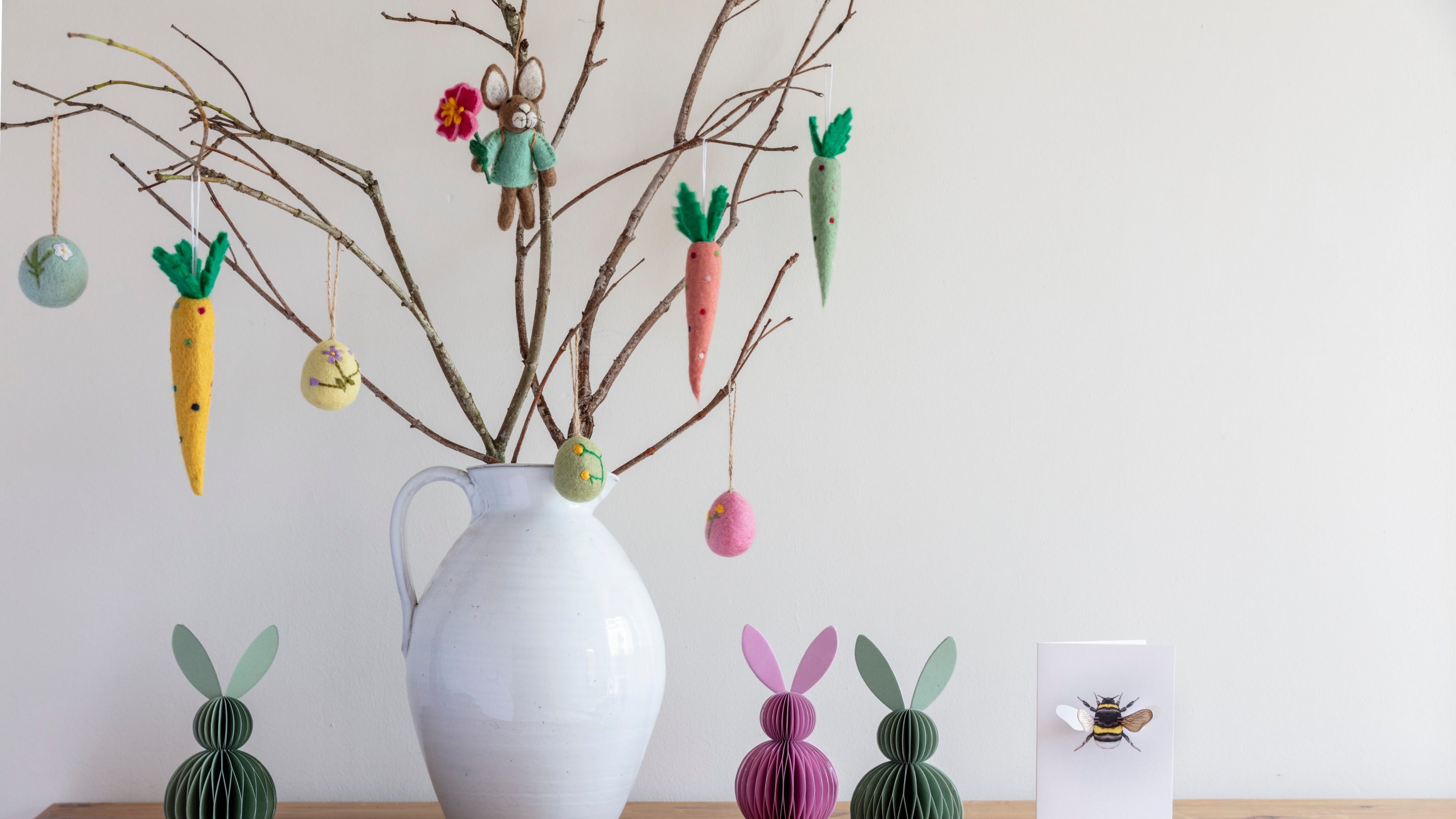 A felt rabbit, eggs and carrots hang as decorations from a vase