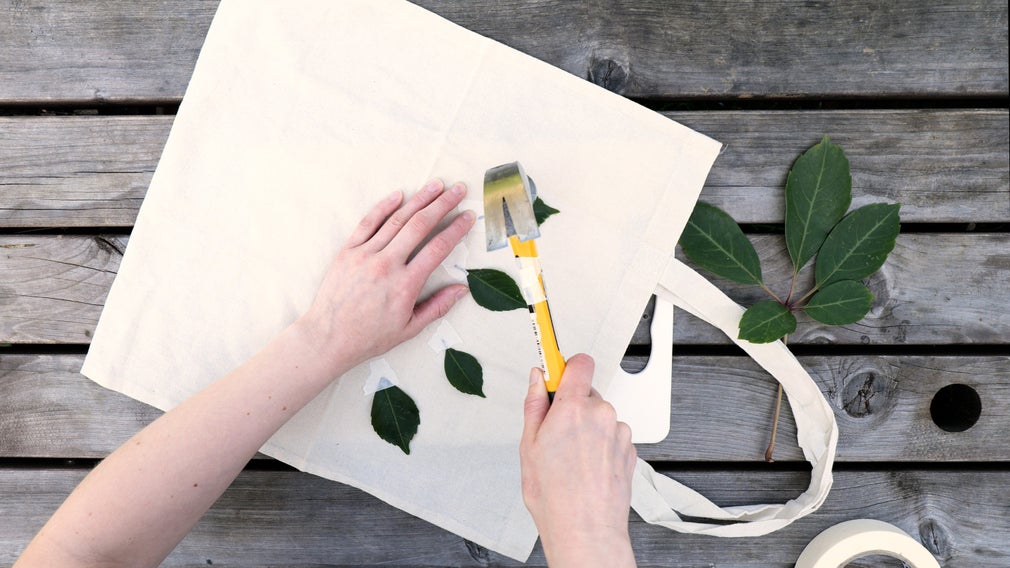 Looking down onto a table where someone is hammering leaves onto a cotton tote bag to create a pattern.