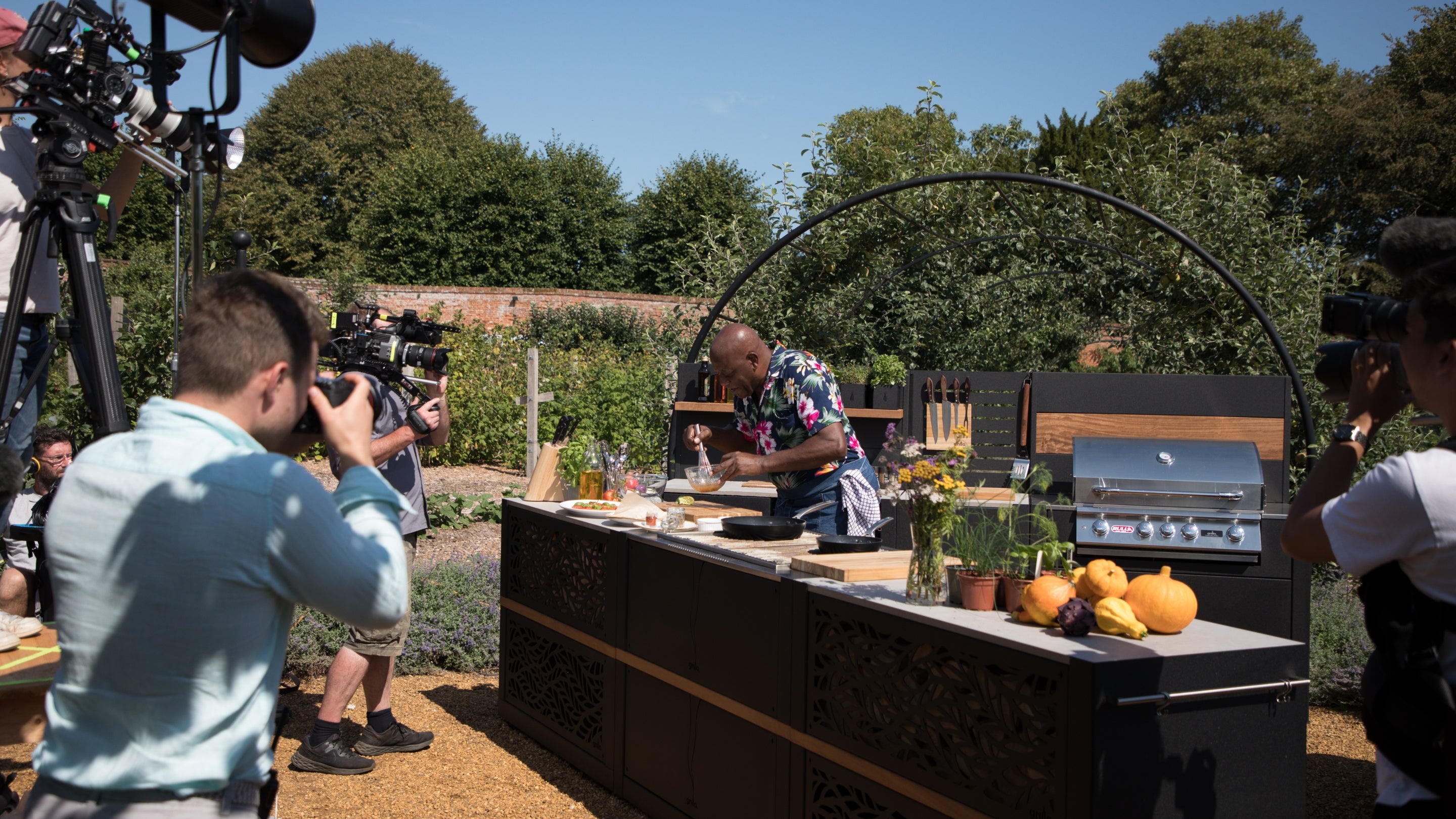 A film crew gathers around an outdoor kitchen set up where Ainsley Harriott is whisking ingredients