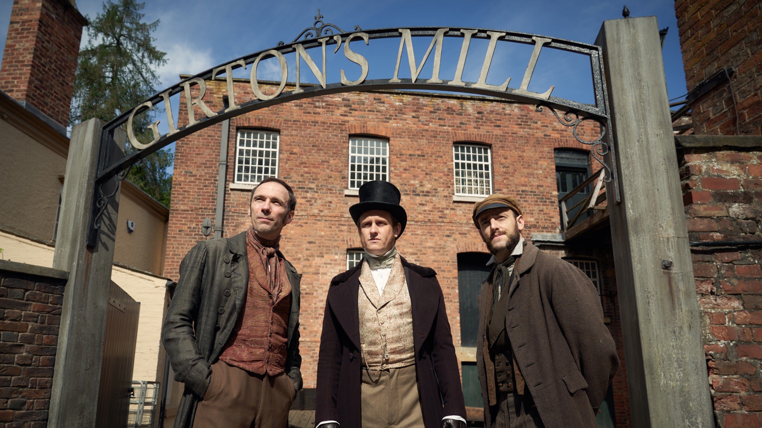 Three men in period costume stand under an entrance arch proclaiming 'Girton's Mill' with the redbrick mill building behind them