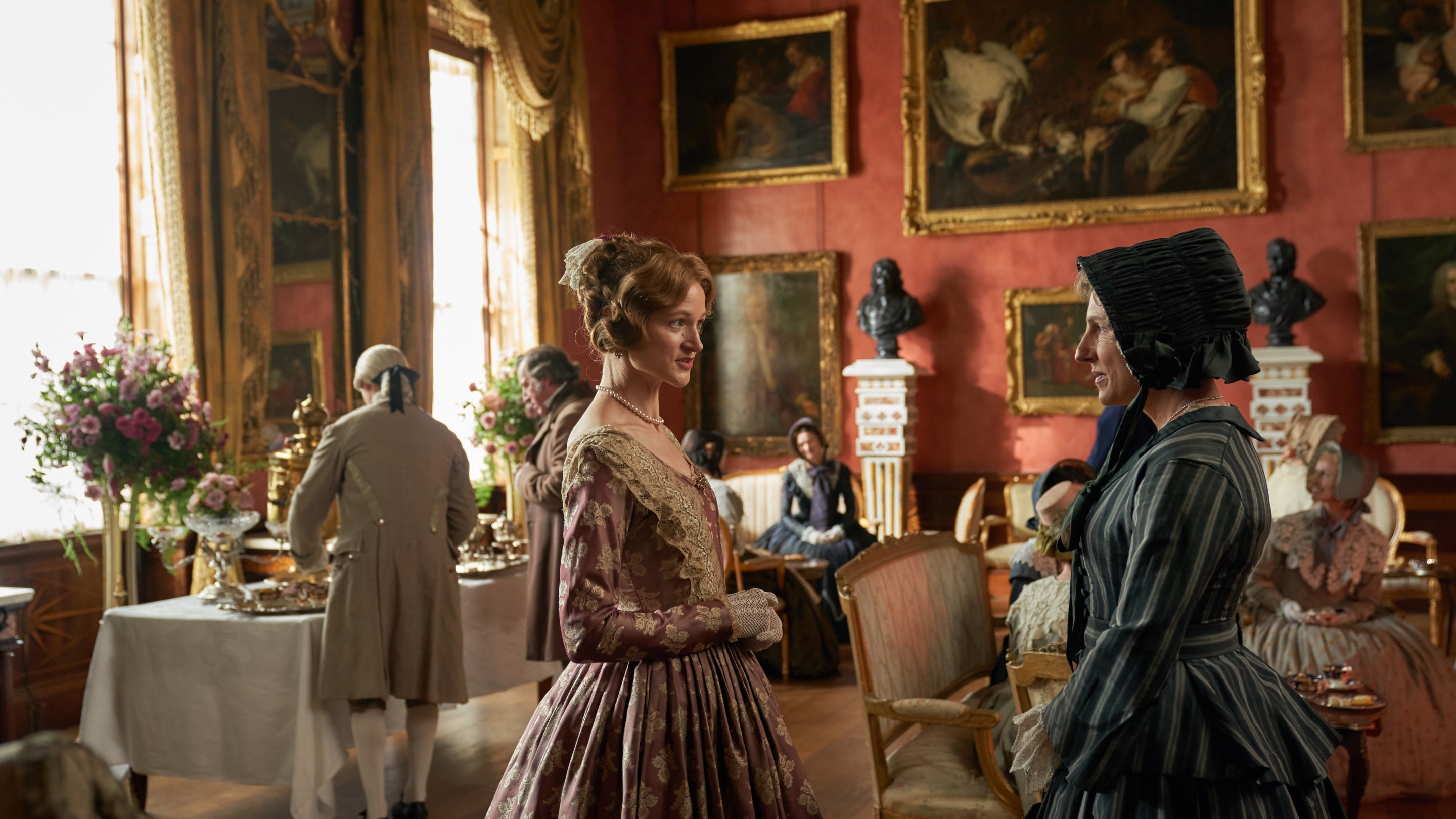 Two women in period costume stand and talk in a grand drawing room