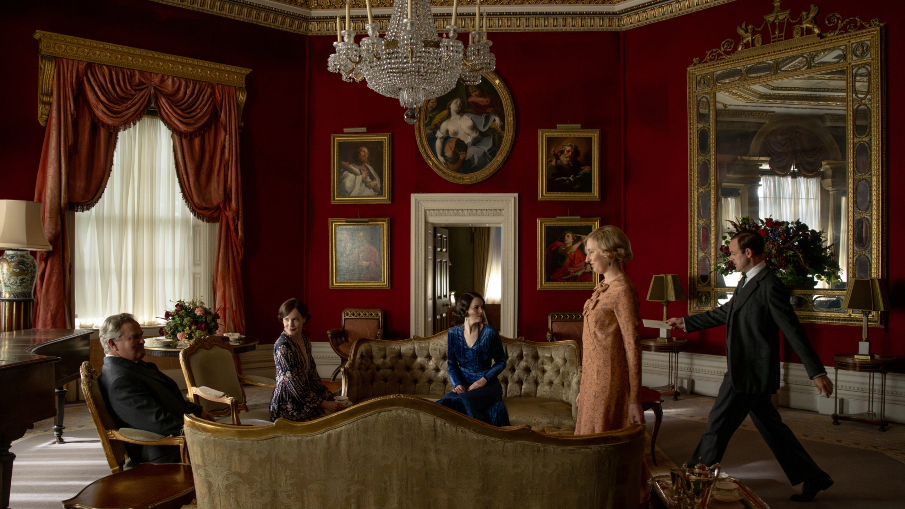 Some of the cast of Downton Abbey sitting on sofas in a circle with two members walking to join them in an ornate room, with red walls and many paintings around them.