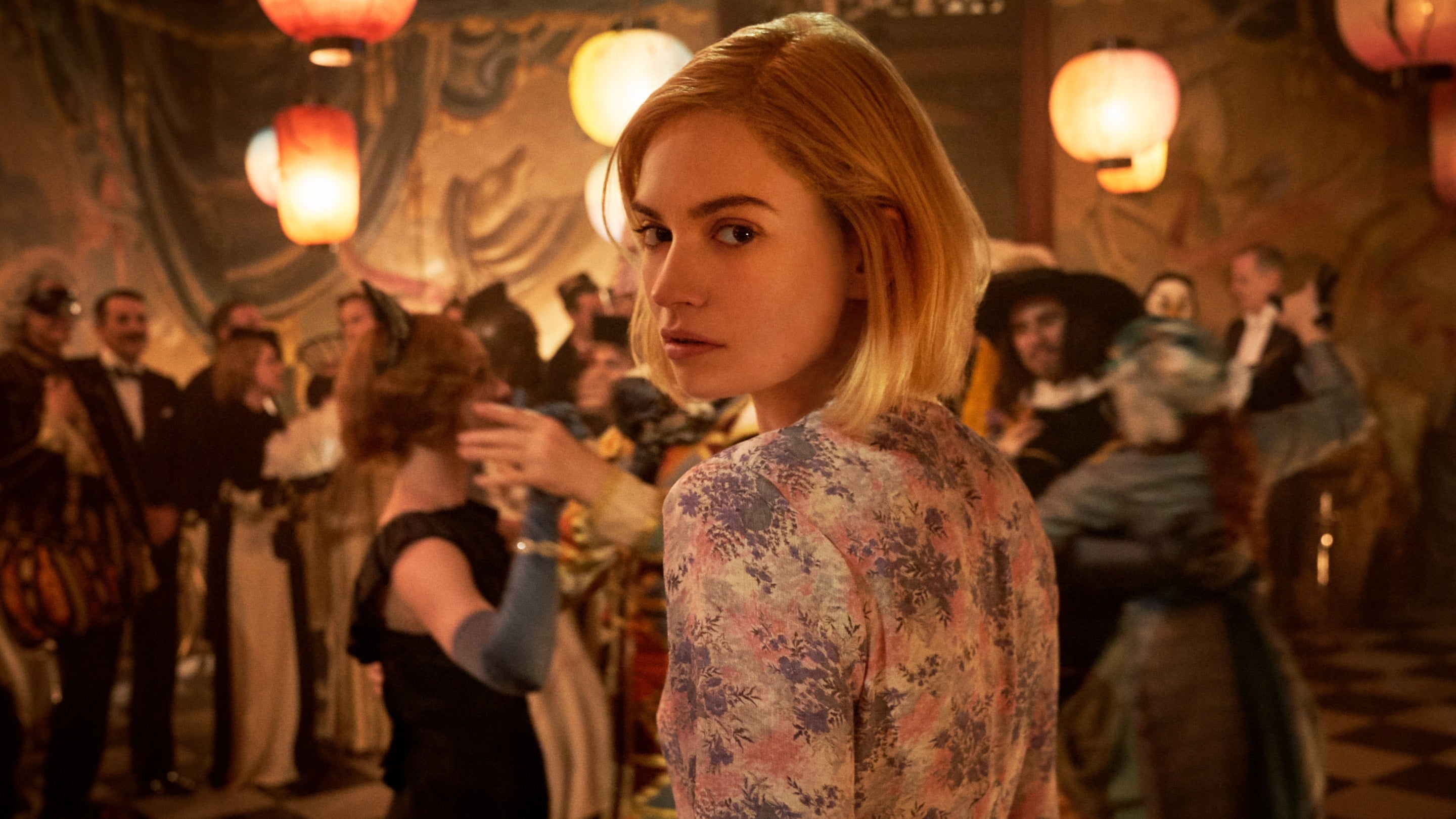 Lily James in the film Rebecca looks at the camera, with costume ball revellers behind her