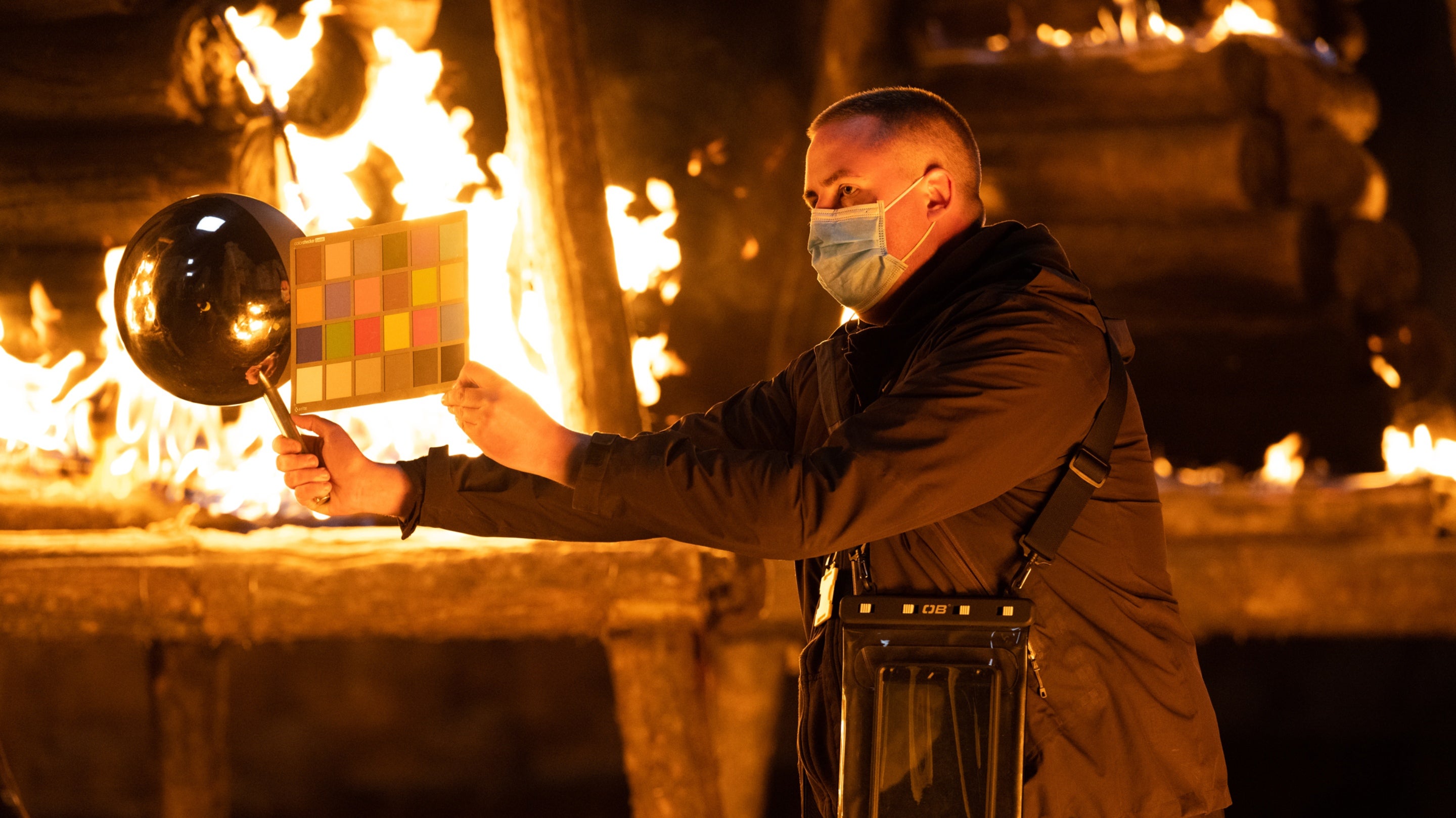 A technician wearing a surgical face mask holds up a large silver ball and a lighting card. There are flames in the background.