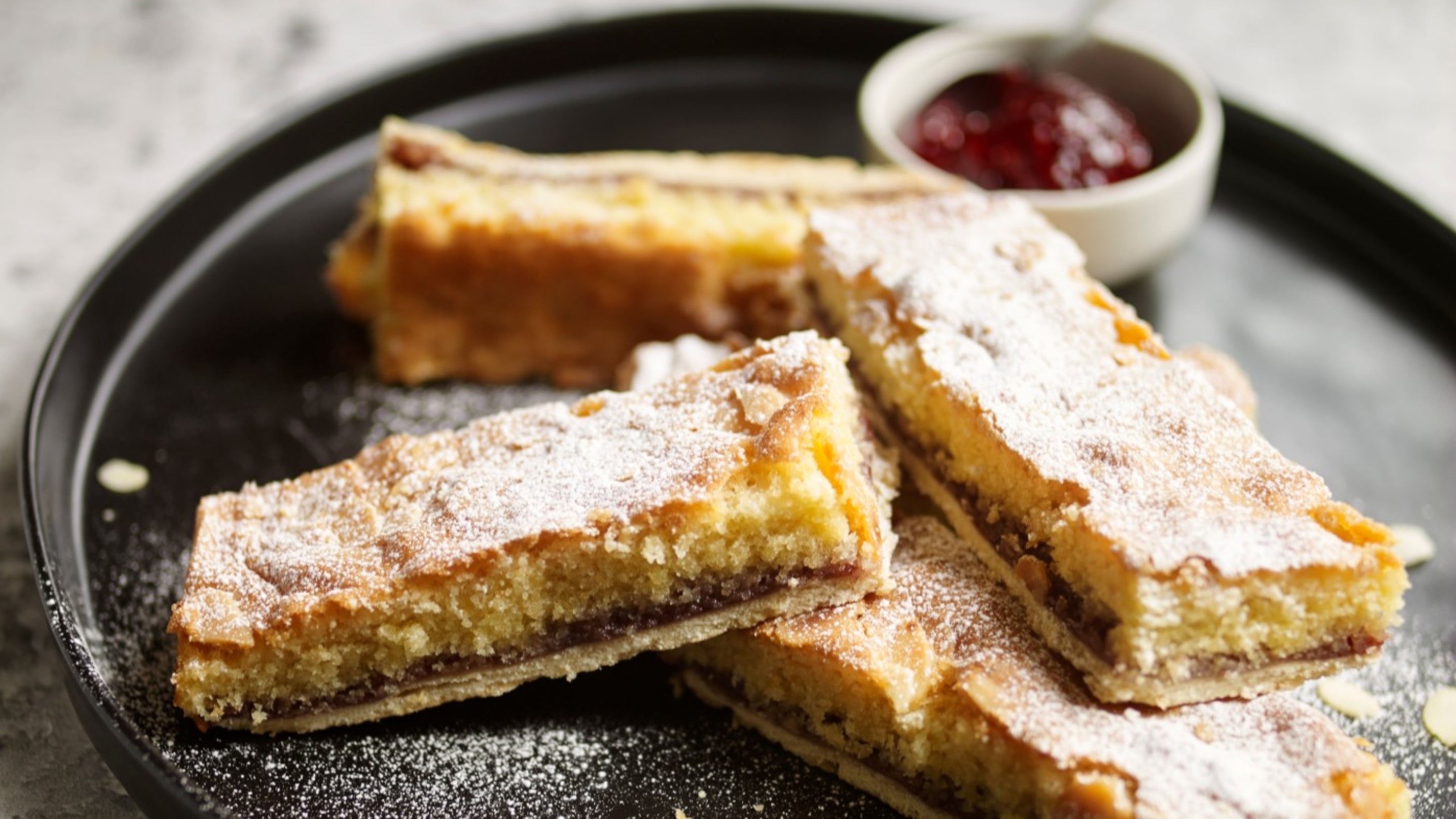 Plate of pre-cut Bakewell slices with pot of raspberry jam to dip in