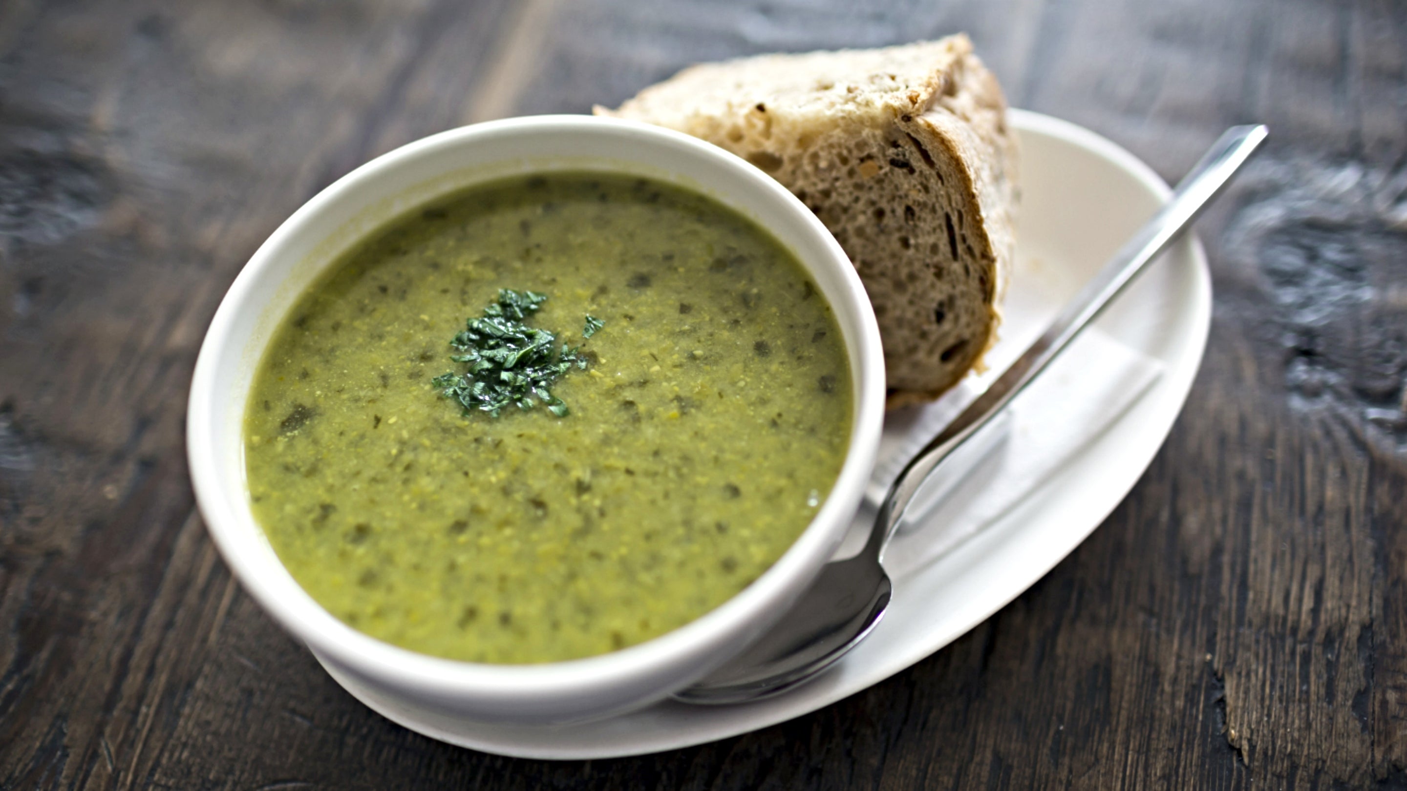 A bowl of leek and potato soup, topped with a sage leaf and oil, with a chunk of brown bread and a spoon on the plate beside it