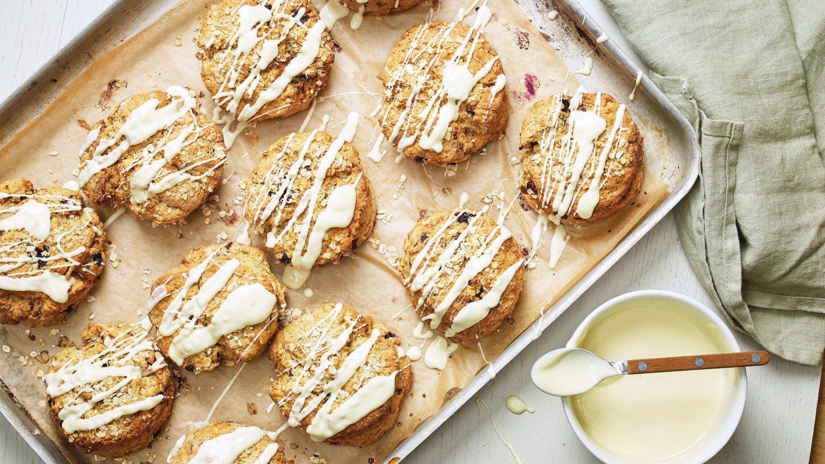 Blueberry and oat scones on a lined baking tray next to a bowl of melted white chocolate and pale green tea towel