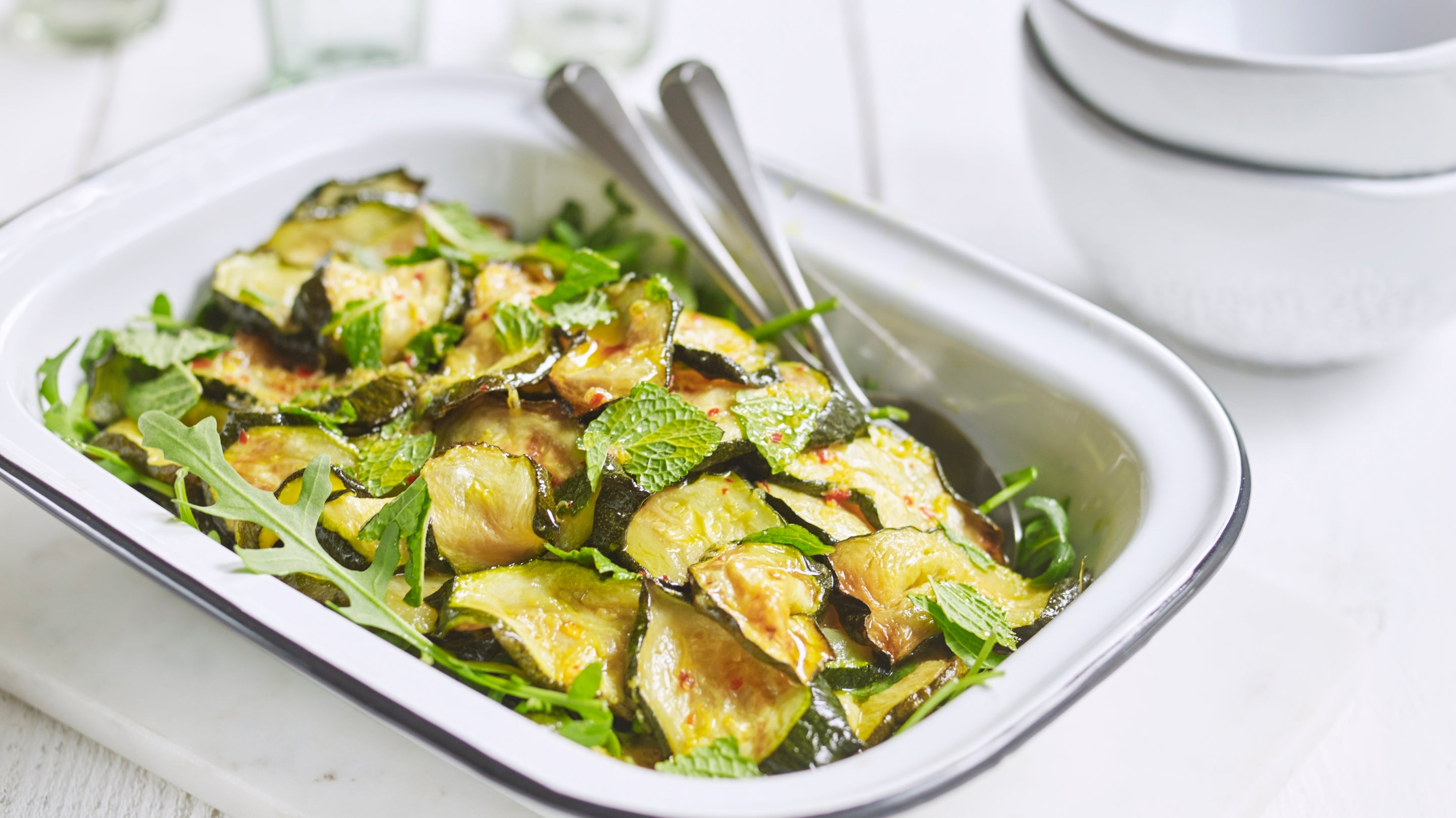 In a rectangular dish, with two serving spoons, are crisp slices of roasted courgette, scattered with fresh mint and rocket