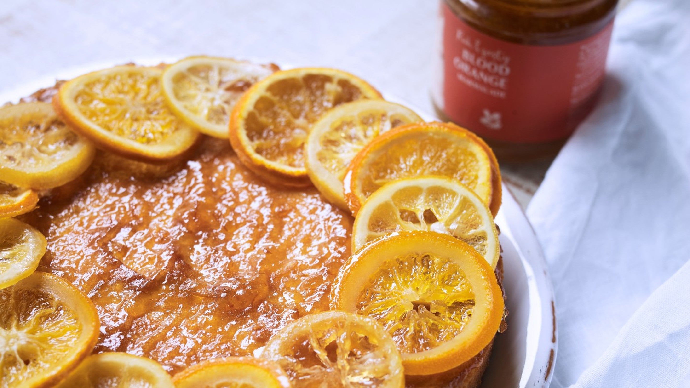 The whole orange and lemon cake on a white plate with a jar of marmalade in the background