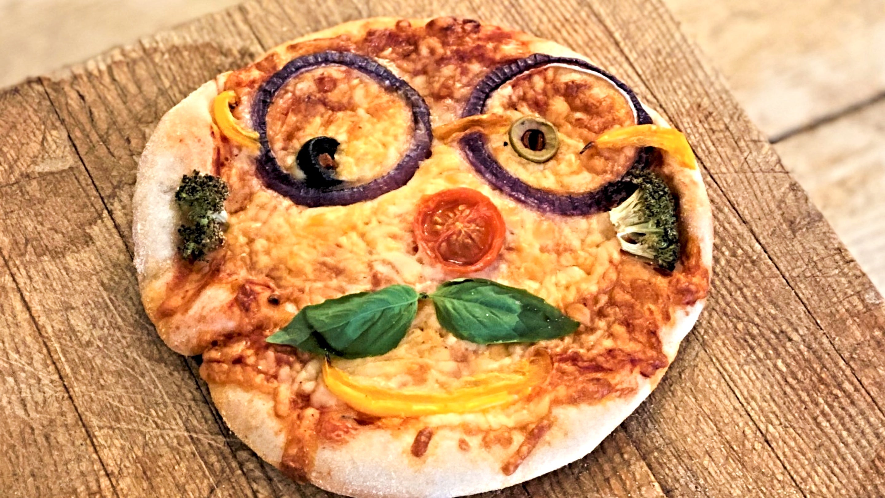 Pizza with a face including olives for eyes, onion rings for glasses, a tomato slice for a nose, two basil leaves for a moustache and broccoli pieces for sideburns
