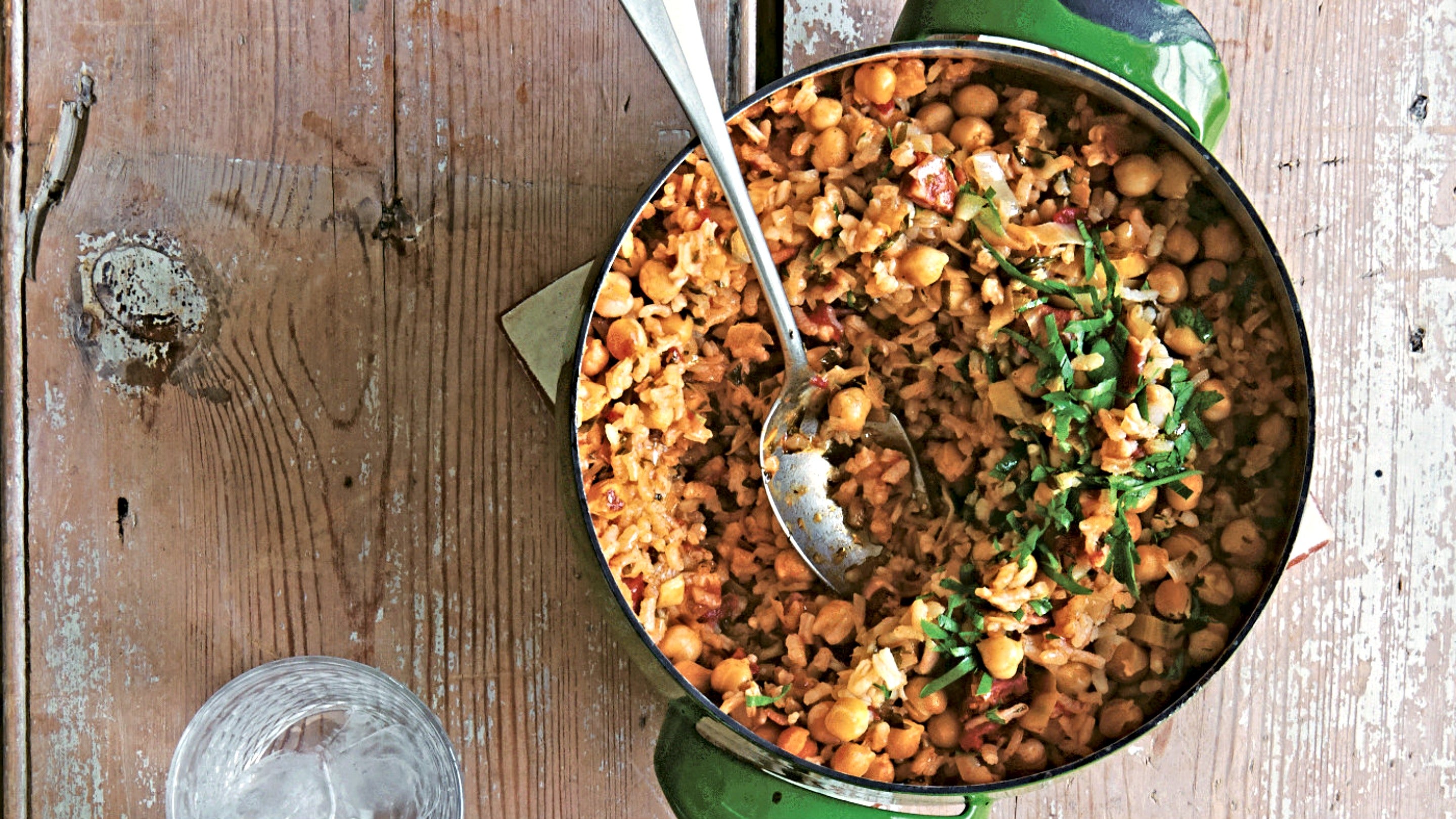A green casserole dish full of Spanish baked rice with leek, bacon and chickpeas, with a spoon in it, on a wooden table