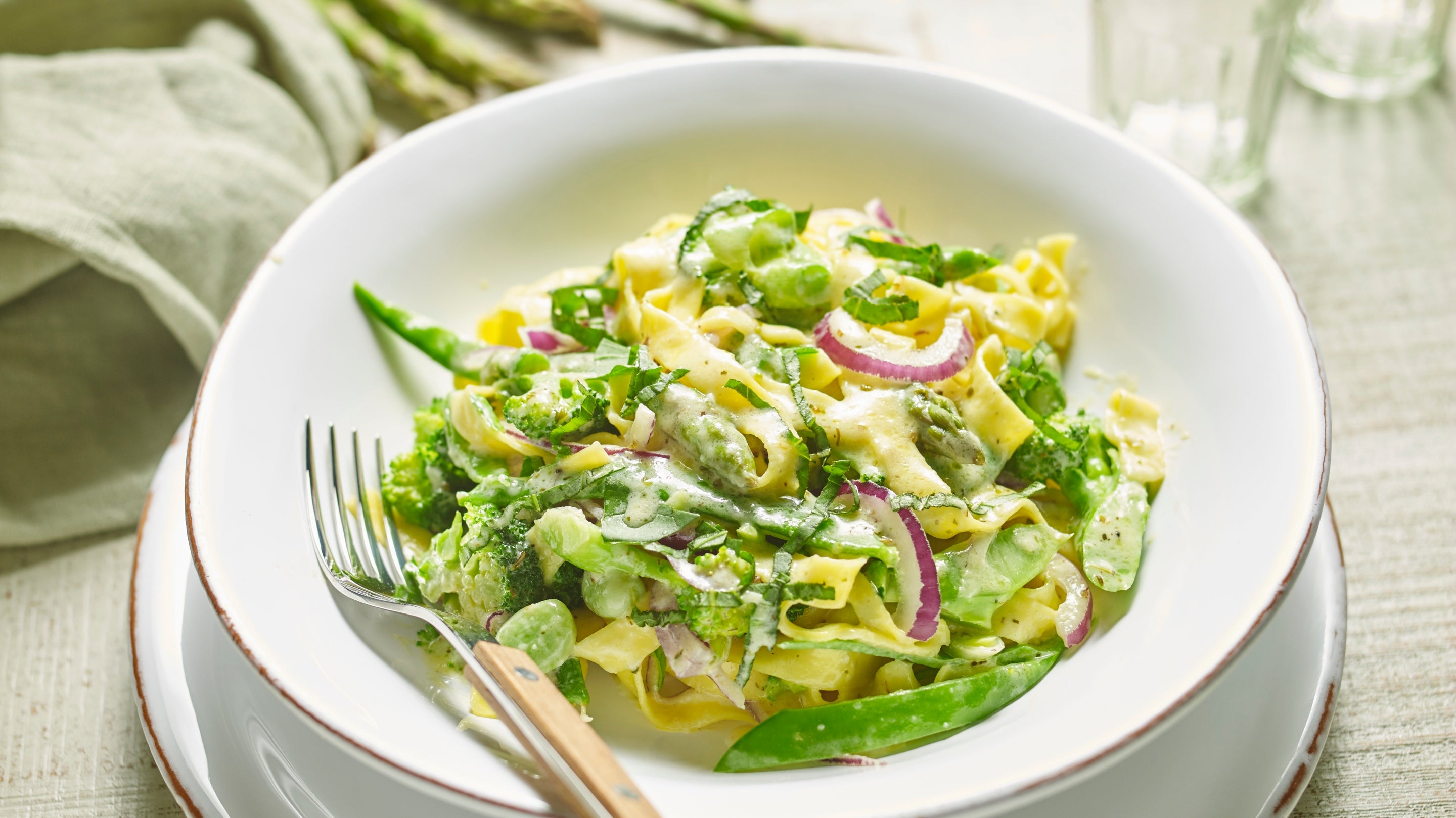 A shallow white bowl with a fork, full of tagliatelle with a creamy vegetable sauce including mange tout and sliced red onion