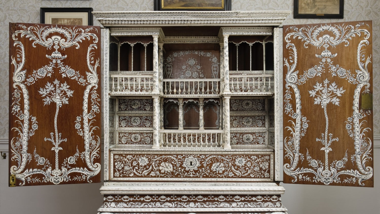 A dark wood cabinet, inlaid with ivory in a design of flowers, leaves and scrolls. It is on a stand, with the doors open to show an interior of drawers and columns, also inlaid with ivory.