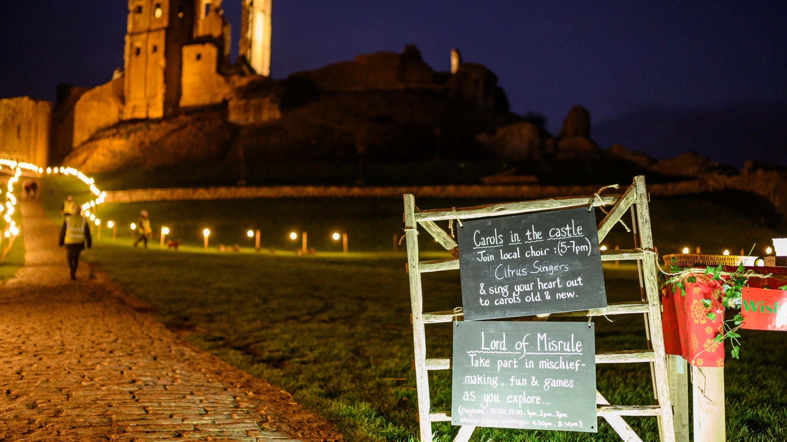 A night time scene of a chalkboard sign on a gate advertising a carol concert next to a path lit by fairy lights that leads to the ruins of Corfe Castle