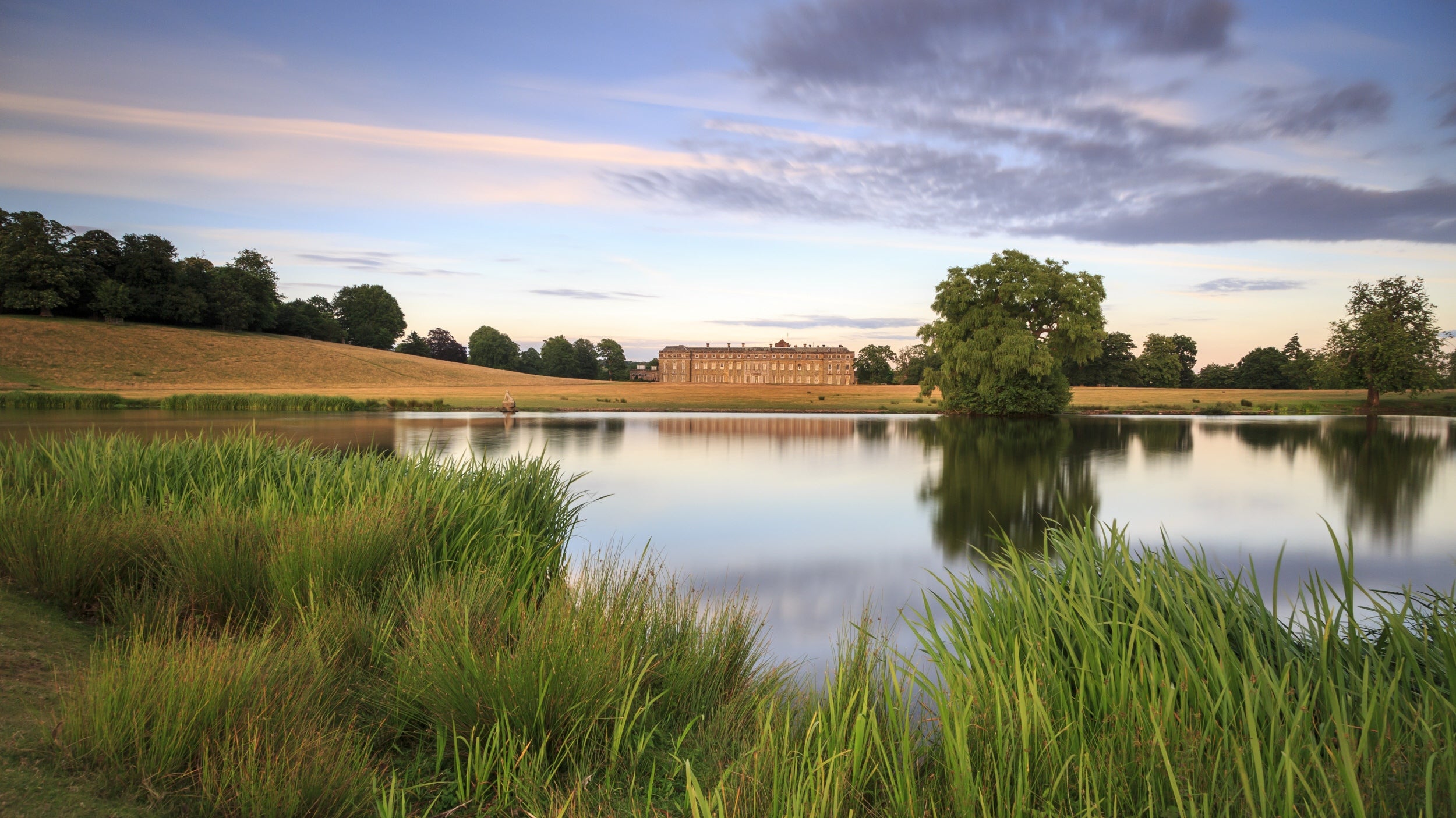 A lake at dawn or dusk with a grand house reflected in it, surrounded by tall green reeds and trees