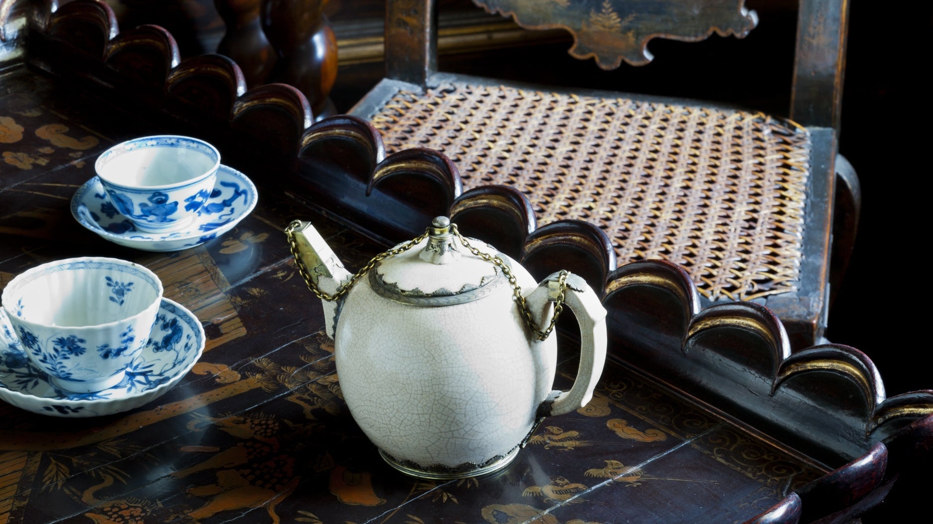 An old Chinese white porcelain teapot set on a a black and brown ornate table with blue willow pattern cups and saucers behind