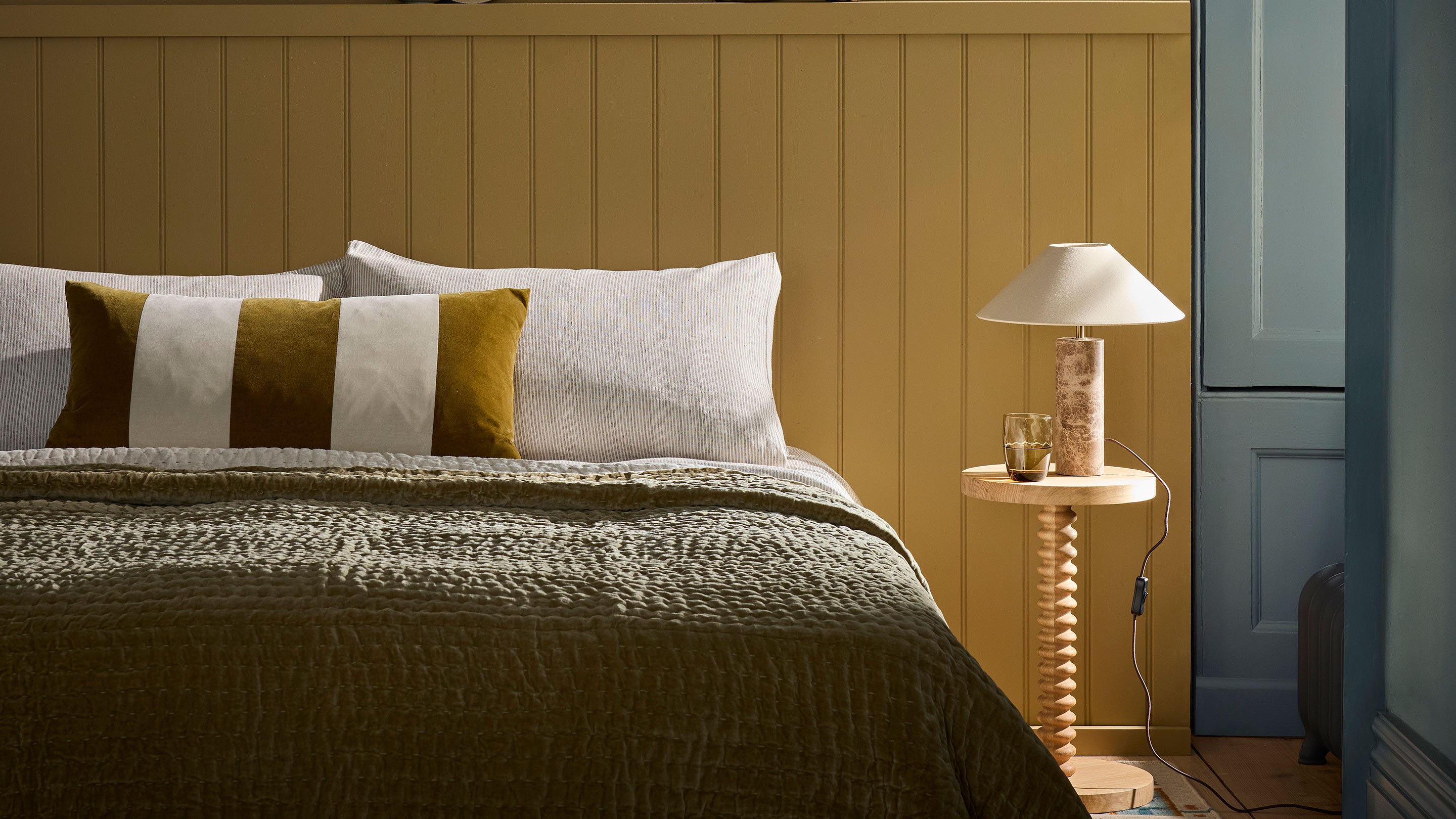 A freshly made bed sits in front of a mustard yellow tongue and groove panelled wall.