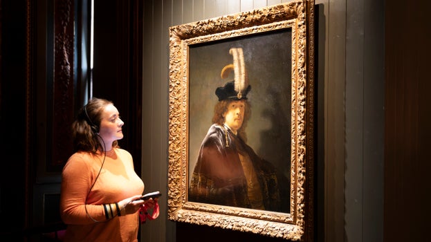 A ldy wearing headphones and holding an audio guide, looks at a painting of a man with a cap with two flambouyant feathers and a large dark velvet cloak with bright silver embroidery on the hem. The man has long curly hair and a thin beard - he is Rembrandt