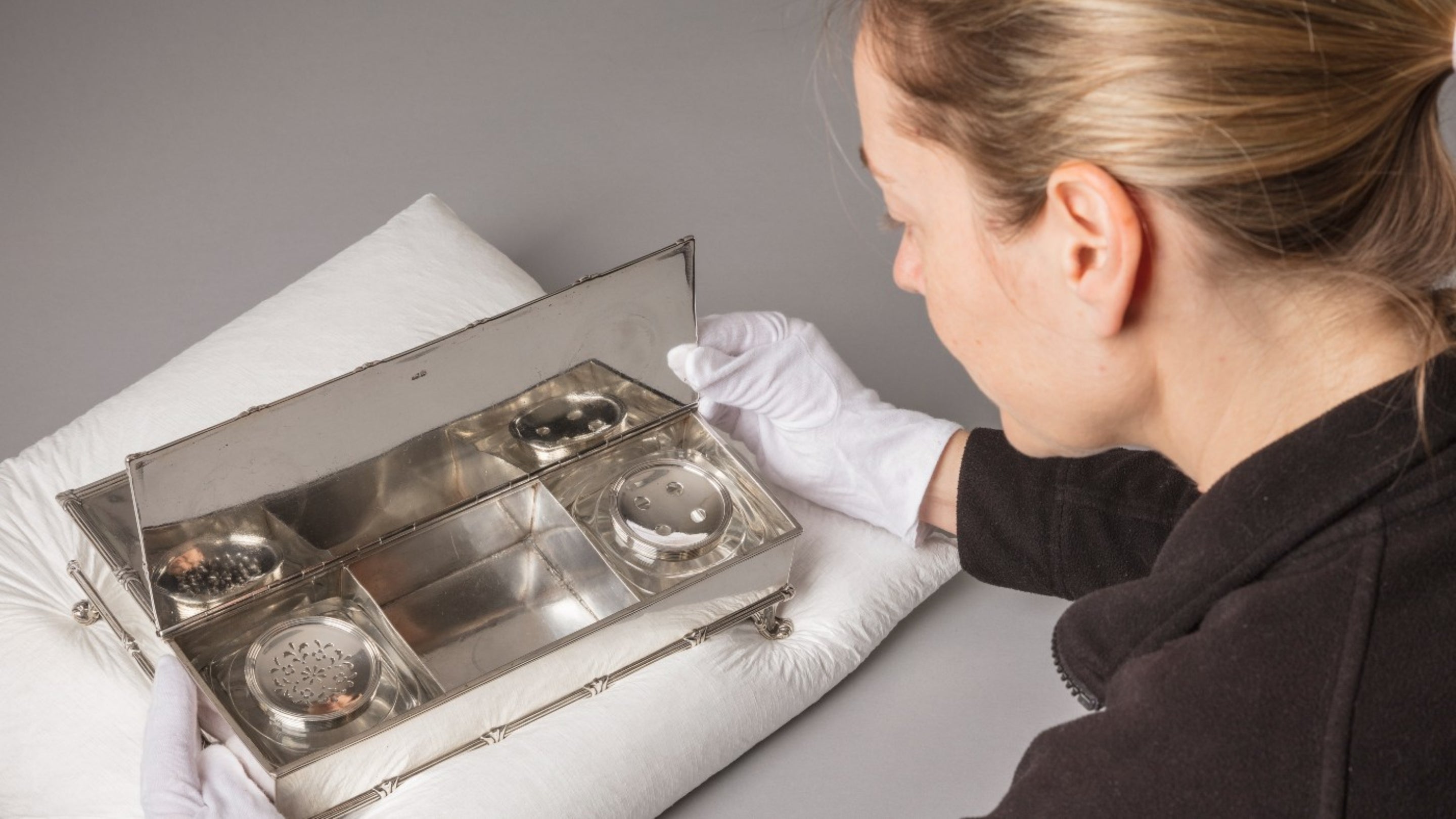 A curator wearing gloves examining a silver inkstand on a pillow