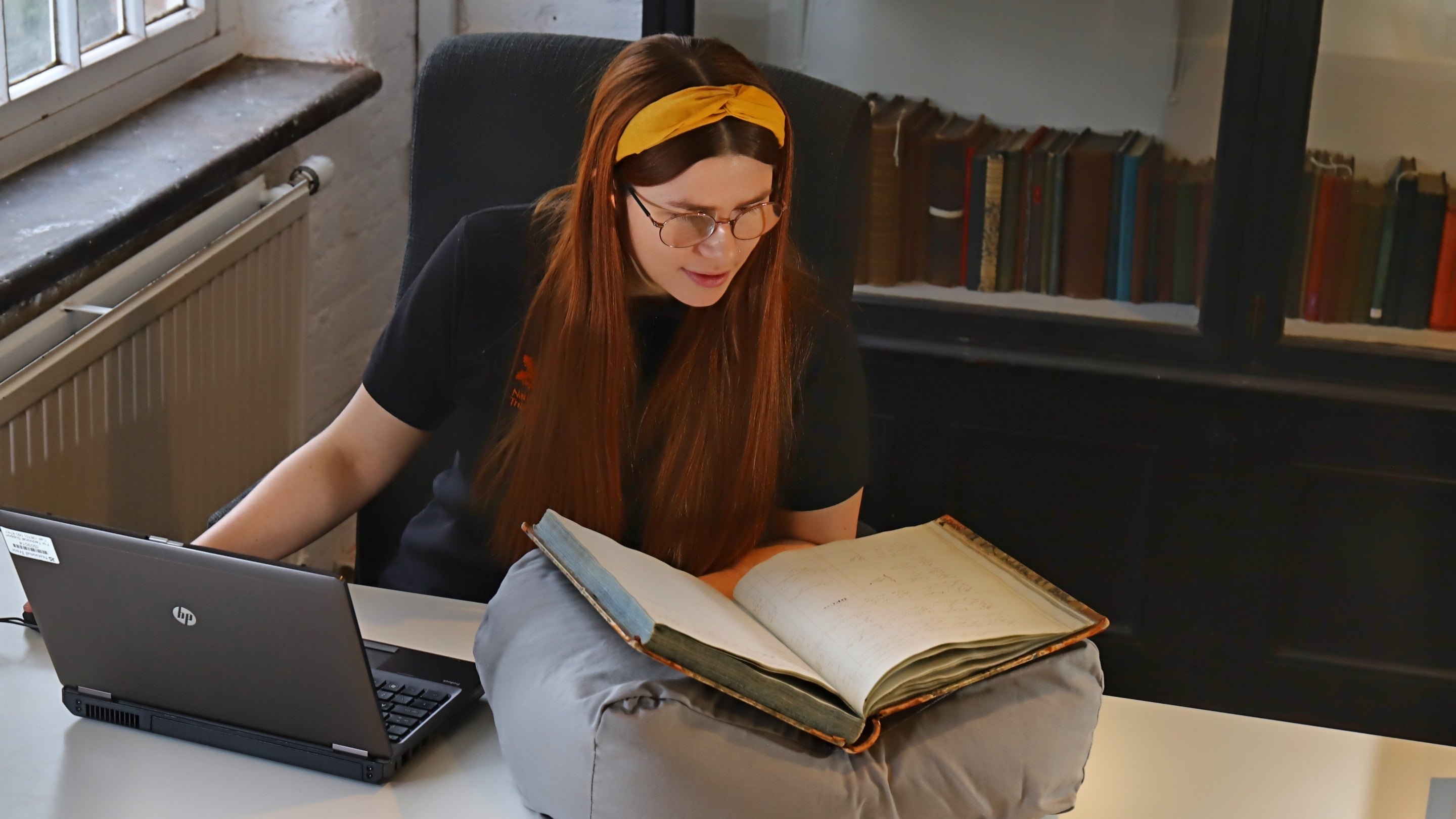 An archivist at Quarry Bank Mill, Cheshire, looks at an old book which is resting on a support cushion and catalogues the information on her laptop.