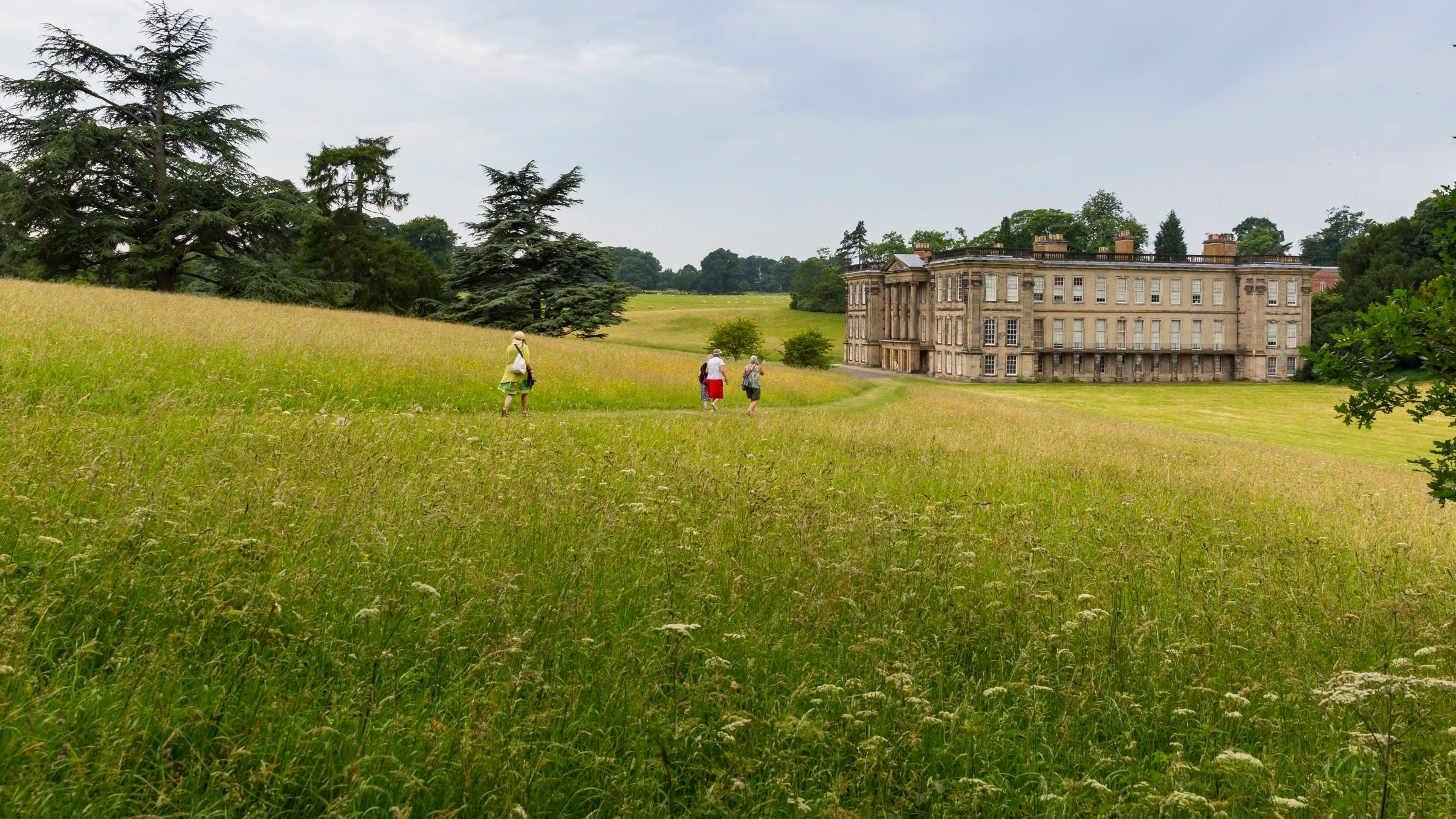 A green grassy meadow with a path through it towards a large, grand building. There are a few people walking along the path in the distance,