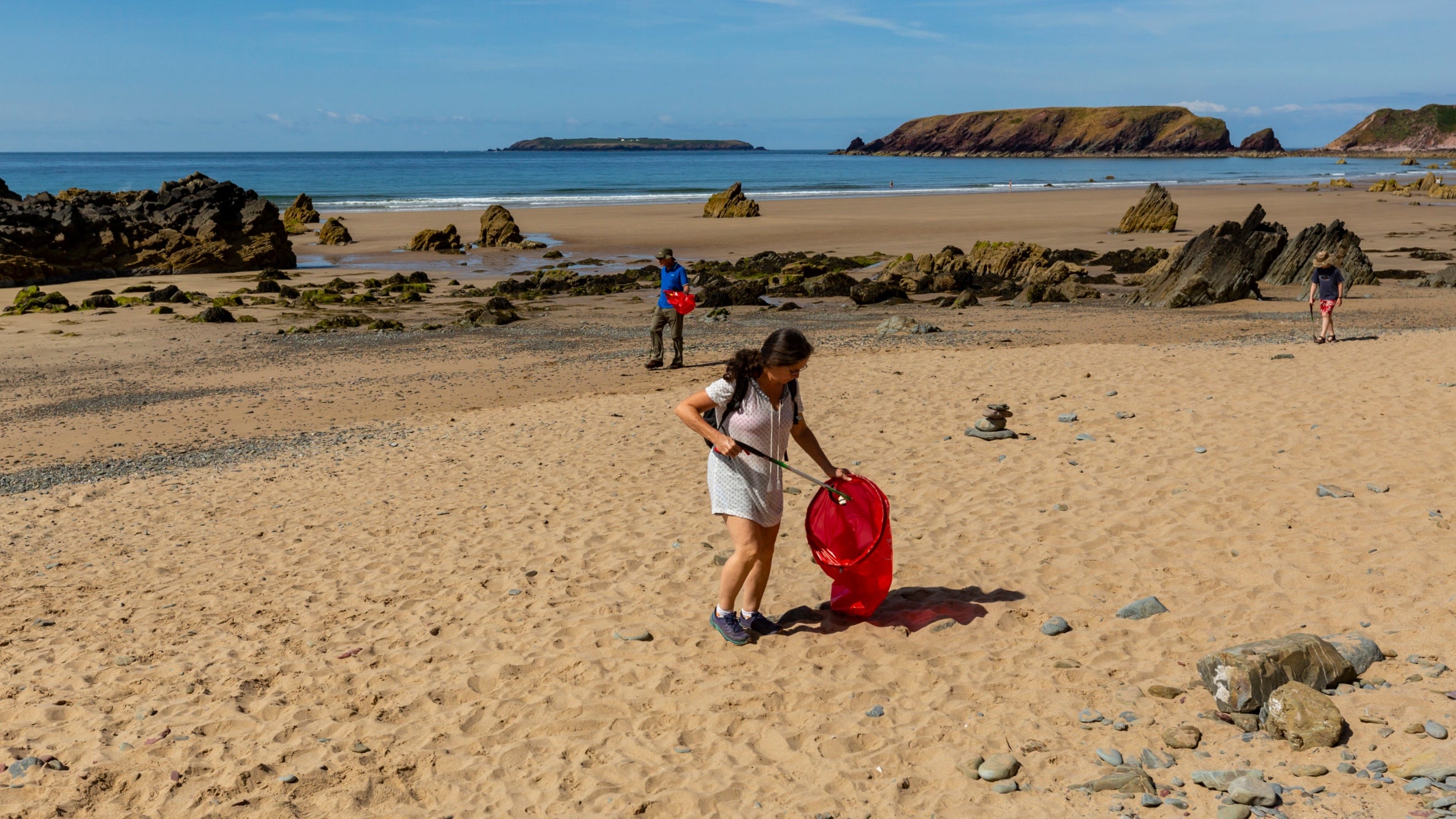A woman in shorts and t-shirt carrying a red sack for rubbish and a litter picker on a sandy beach on a sunny day with the sea behind in the distance.